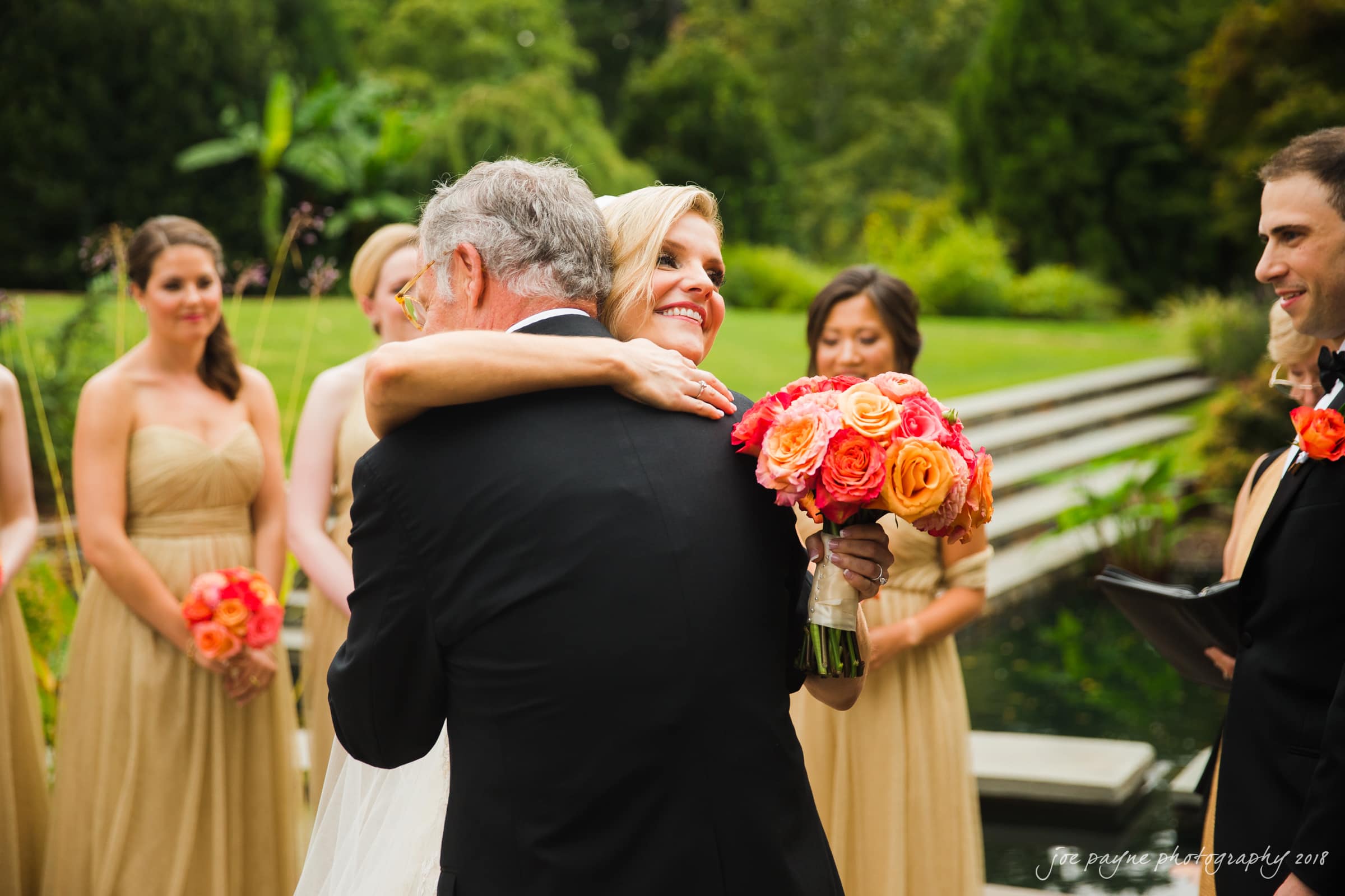 angle amphitheater at duke gardens wedding - katherine & andy A bride holding a vibrant bouquet of orange and pink flowers hugs an older man, possibly her father, during an outdoor wedding ceremony. Bridesmaids in beige dresses and lush greenery surround them.