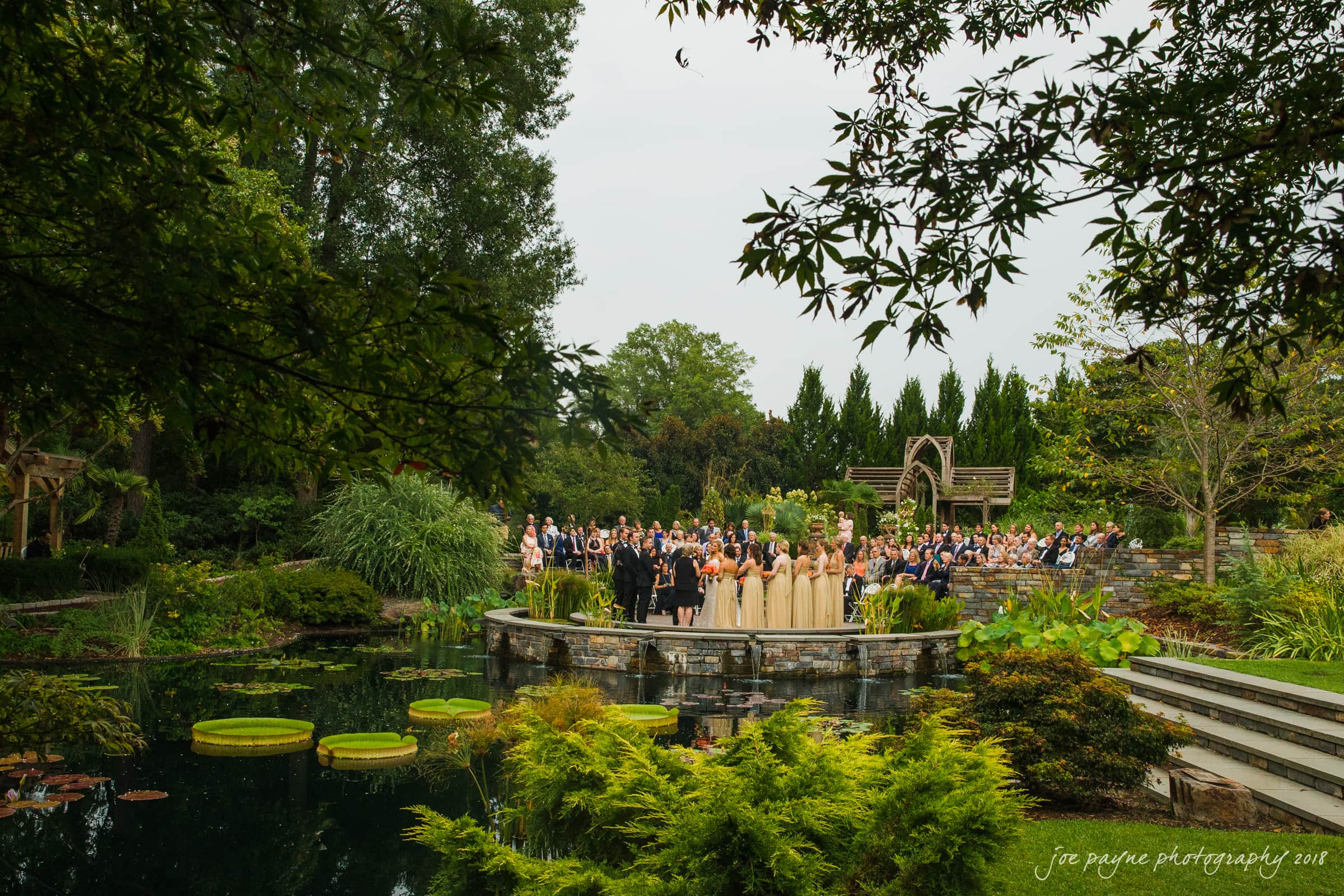 angle amphitheater at duke gardens wedding - katherine & andy A wedding ceremony held in a lush garden setting by a pond. Guests are seated on one side, while the wedding party stands on a stone platform. Greenery surrounds the area, and large lily pads float on the water.