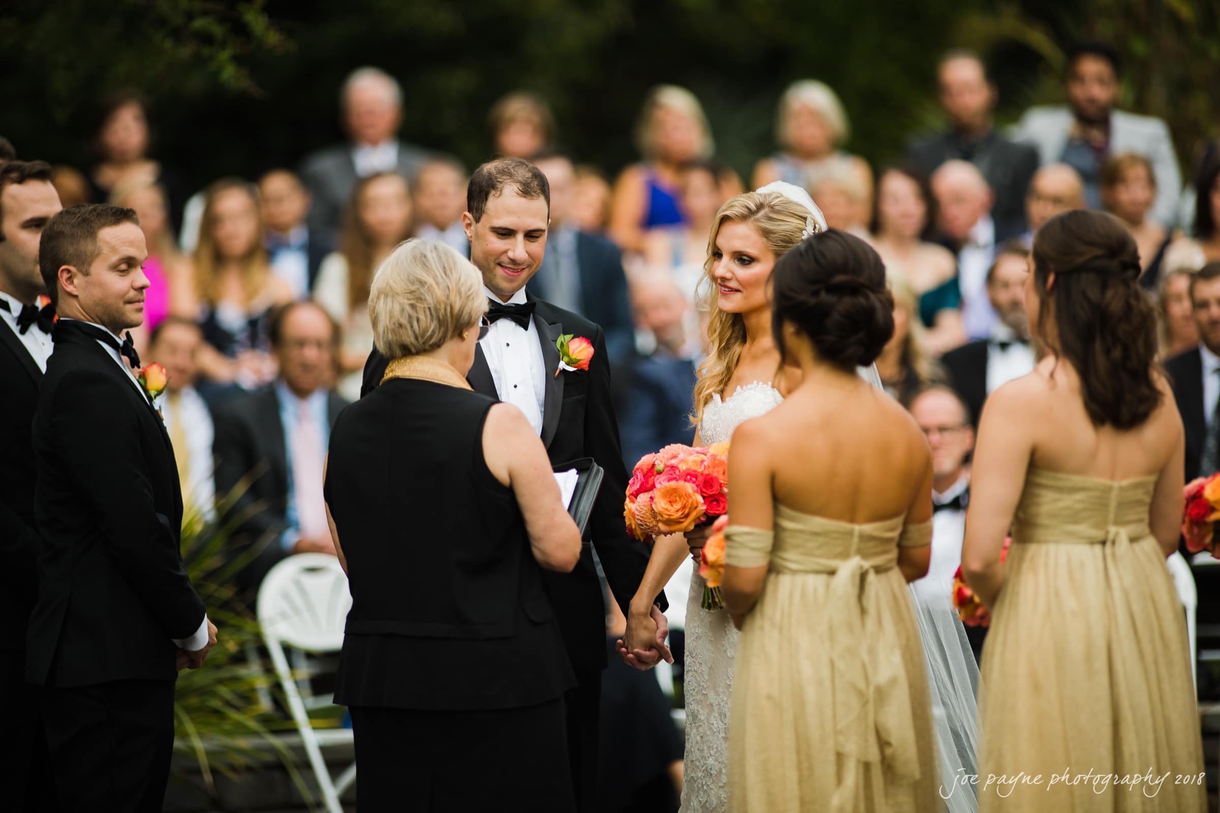 angle amphitheater at duke gardens wedding - katherine & andy A bride and groom stand at an outdoor wedding ceremony, holding hands and facing the officiant. The bride wears a white gown, holding a bouquet of orange flowers. Bridesmaids in gold dresses stand nearby, with guests seated in the background.