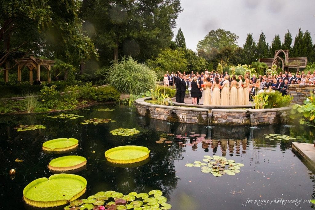 A Duke Gardens wedding unfolds gracefully by a large pond adorned with lily pads. The bride and groom, along with their bridal party, stand elegantly as guests sit across the lush, green setting. Majestic trees and enchanting garden structures provide a picturesque backdrop to this special day.