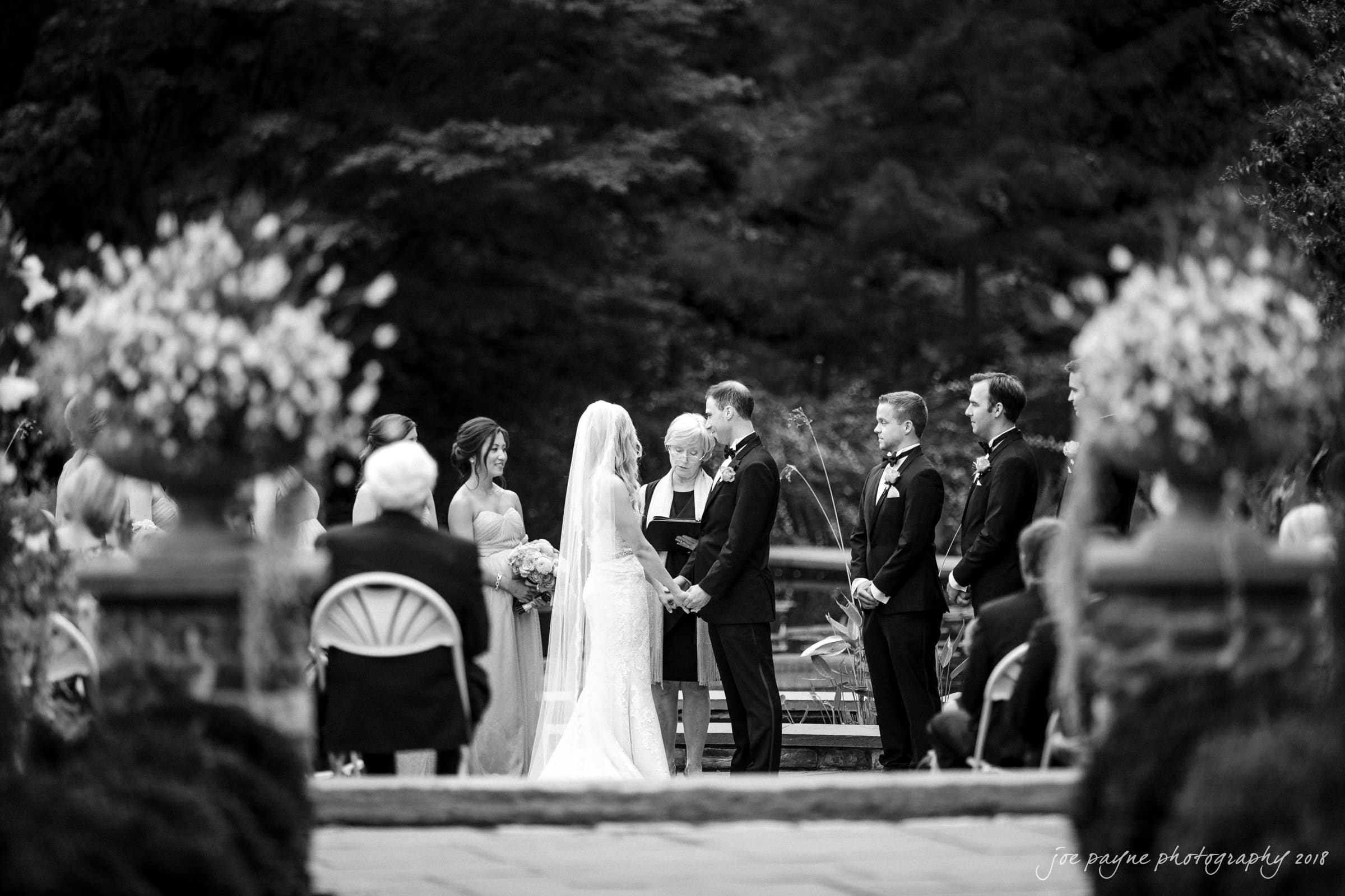 angle amphitheater at duke gardens wedding - katherine & andy A black and white photo capturing a wedding ceremony outdoors. The bride and groom stand at the altar, surrounded by the wedding party and guests. Floral arrangements and trees frame the scene in a formal garden setting.