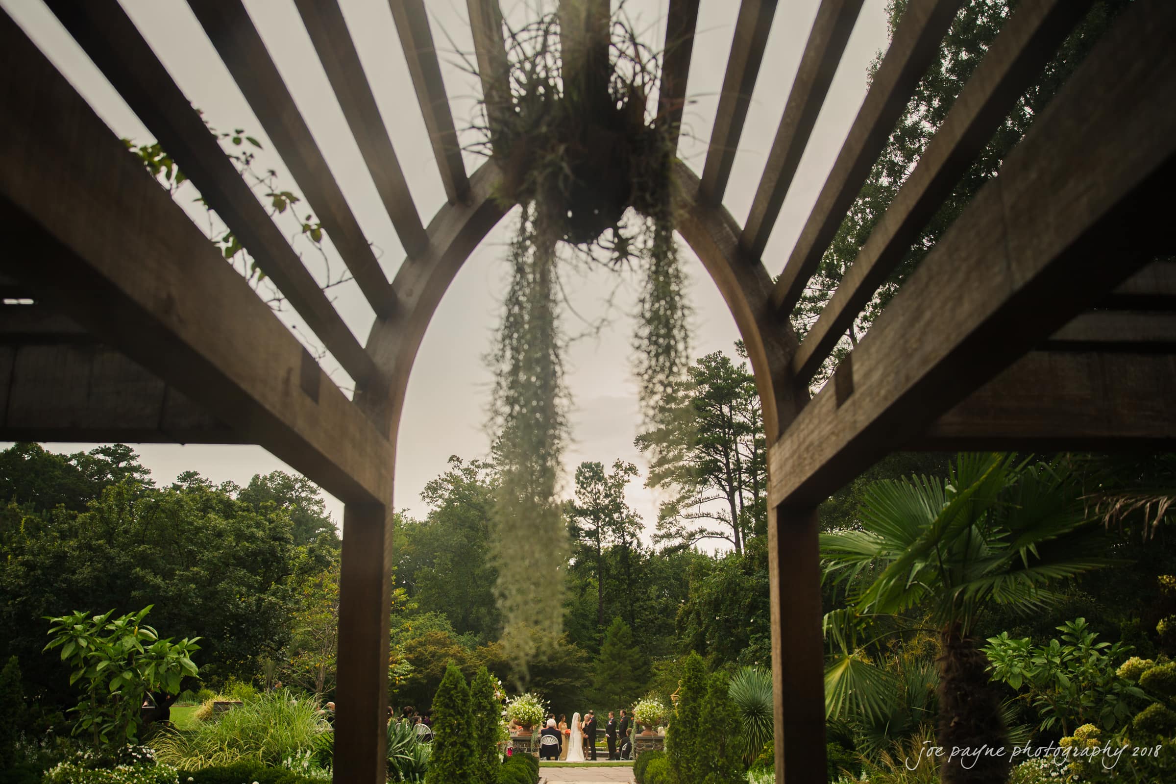 angle amphitheater at duke gardens wedding - katherine & andy A wooden pergola frames a garden with lush greenery and tall trees. A hanging plant is in the center, and a wedding ceremony is visible in the background with guests seated and the couple at the altar.