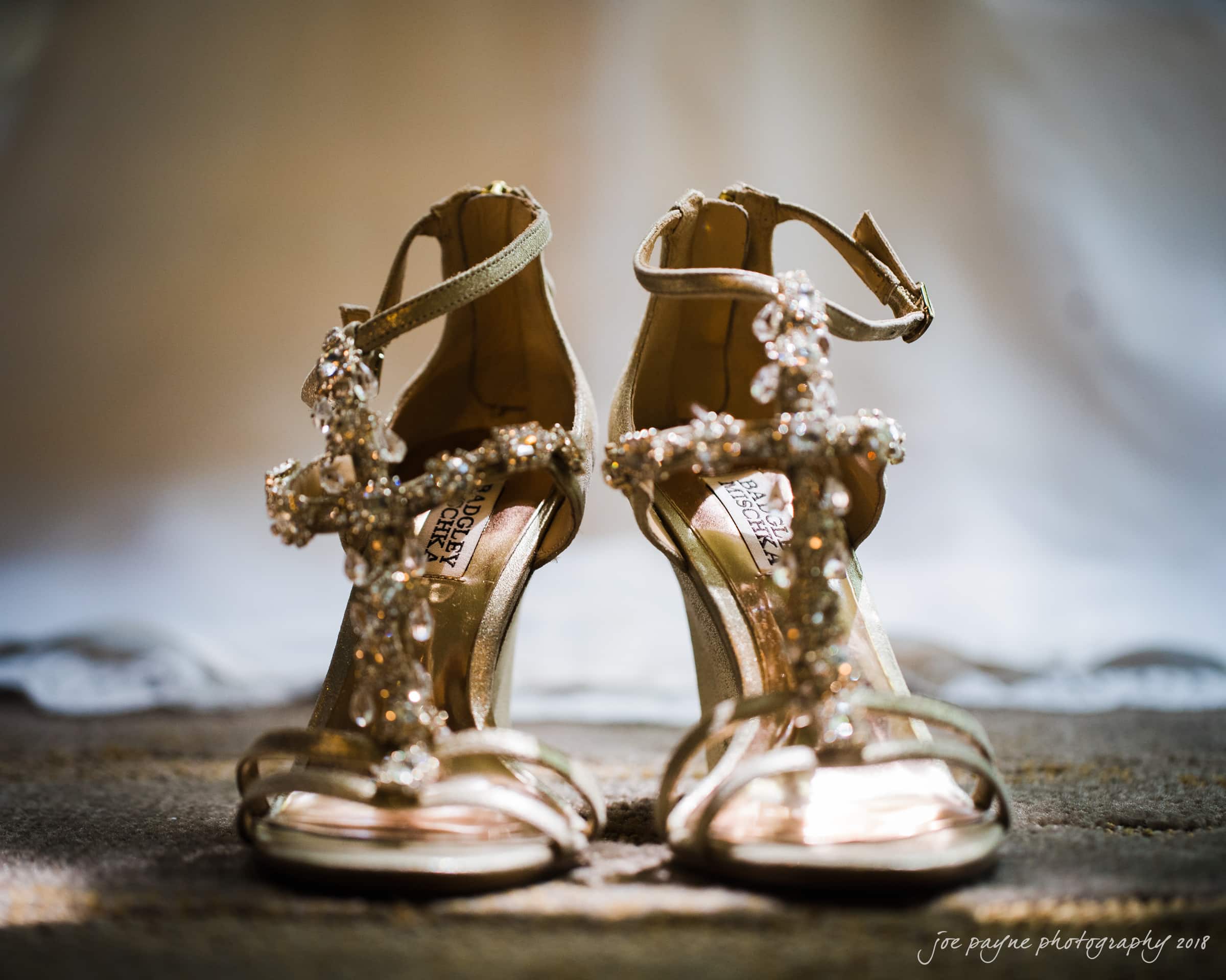 angle amphitheater at duke gardens wedding - katherine & andy A pair of elegant gold high-heeled sandals adorned with intricate beading and crystal embellishments, displayed on a soft, textured surface. The background is softly blurred, creating a warm, intimate atmosphere.