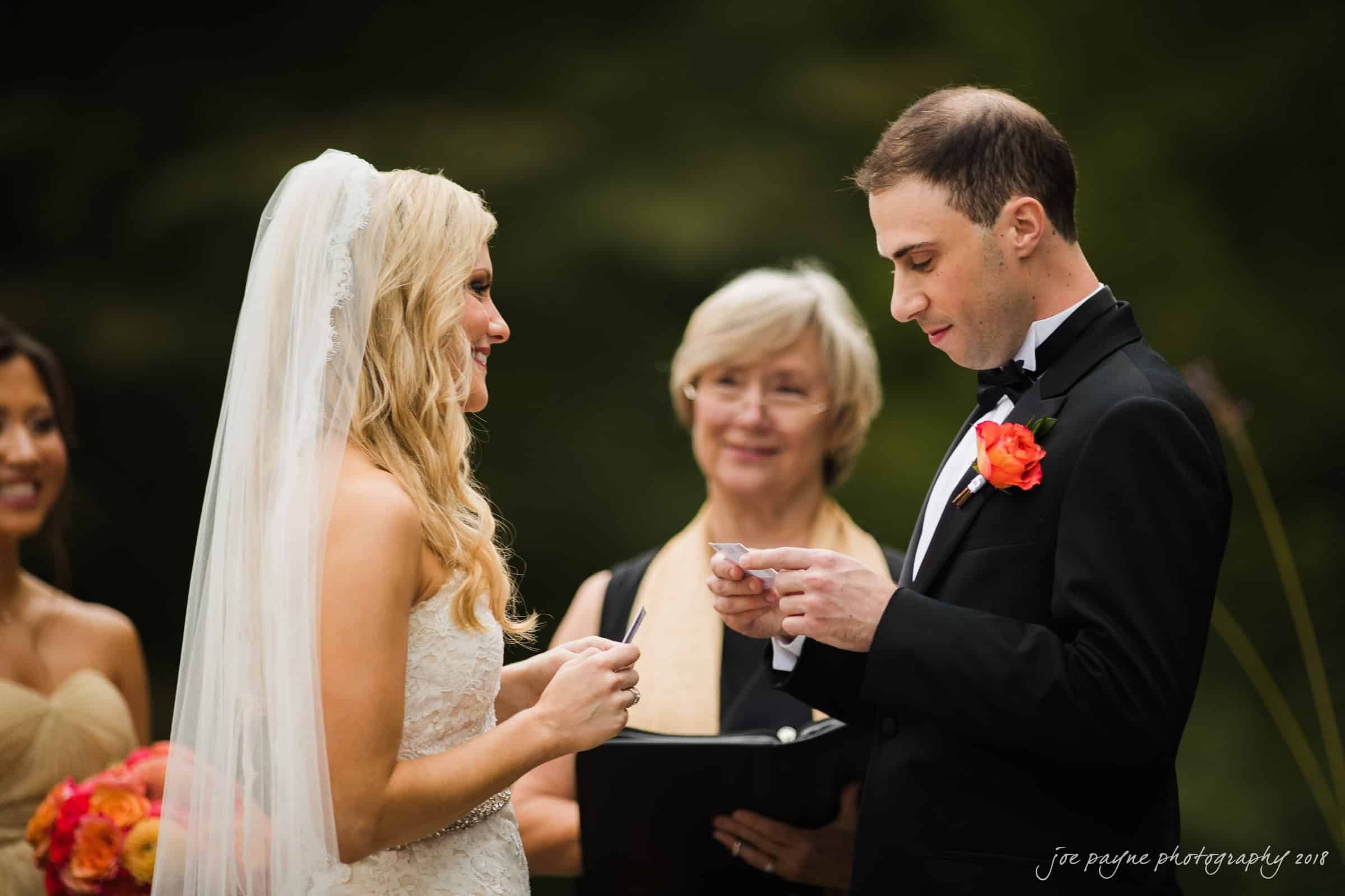 angle amphitheater at duke gardens wedding - katherine & andy A bride and groom stand facing each other during a wedding ceremony. The bride holds a card and wears a white gown and veil. The groom, in a black suit with a red boutonniere, reads from a card. An officiant stands between them, smiling.