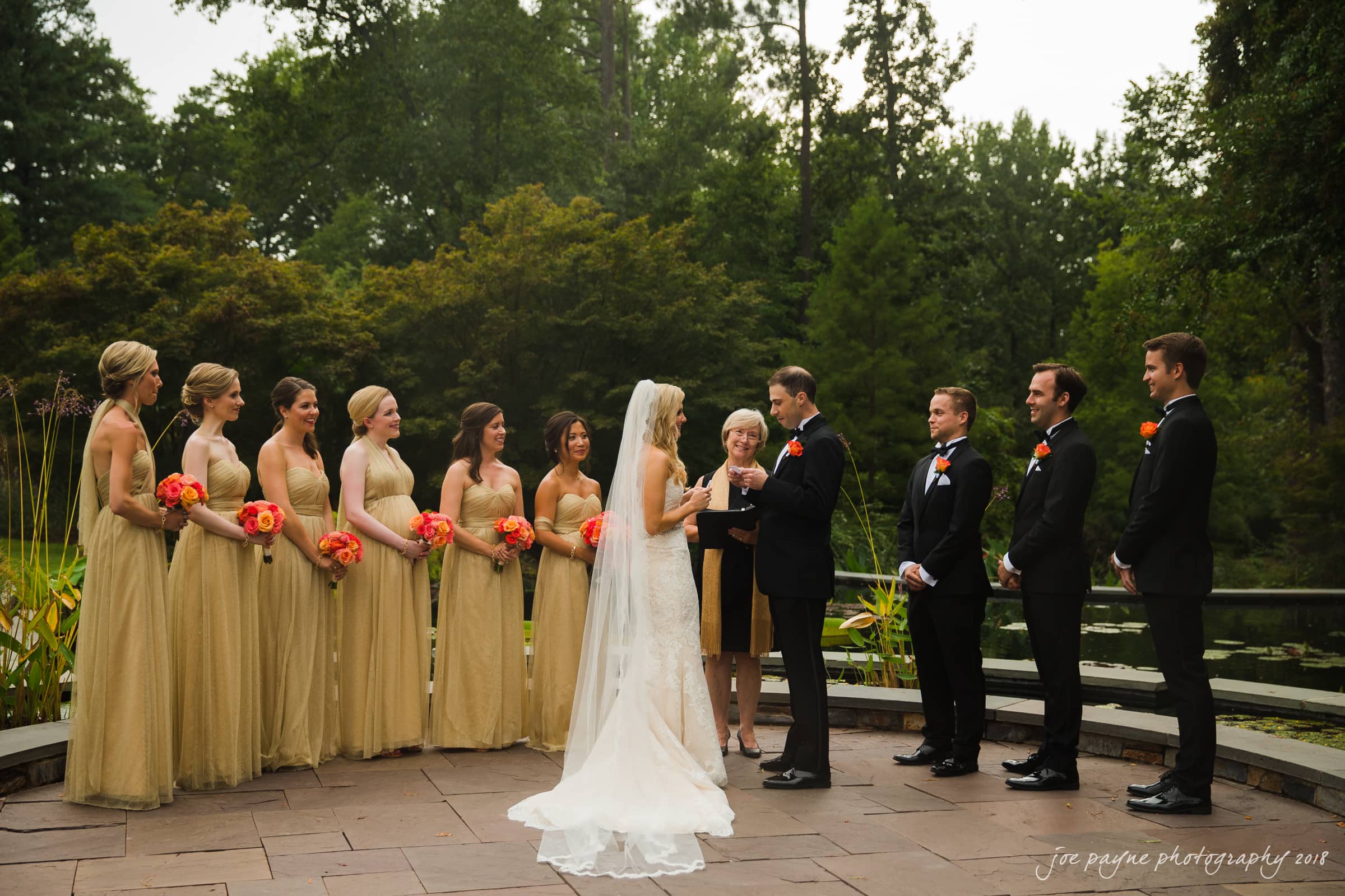 angle amphitheater at duke gardens wedding - katherine & andy A bride and groom exchange vows outdoors with a bridal party. The bride wears a white dress, and bridesmaids in gold hold orange flowers. Groomsmen in black suits stand beside the groom. Lush greenery surrounds the scene.