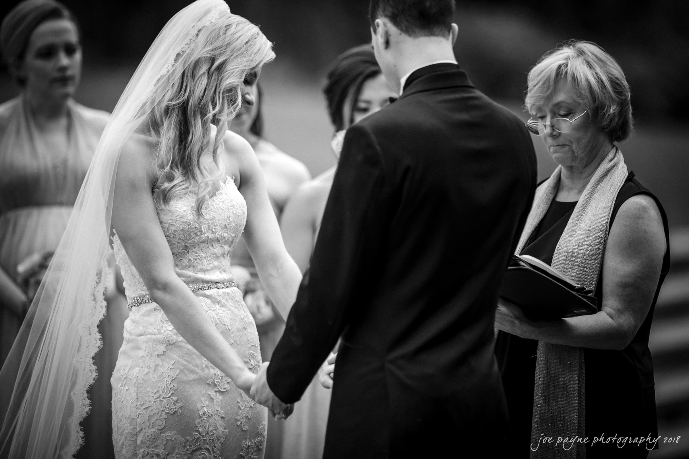 angle amphitheater at duke gardens wedding - katherine & andy A bride and groom hold hands during a wedding ceremony. The bride wears a lace gown and veil, while the groom is in a suit. A woman, possibly an officiant, stands nearby with a book. Bridesmaids are in the background. The scene is emotional and intimate.