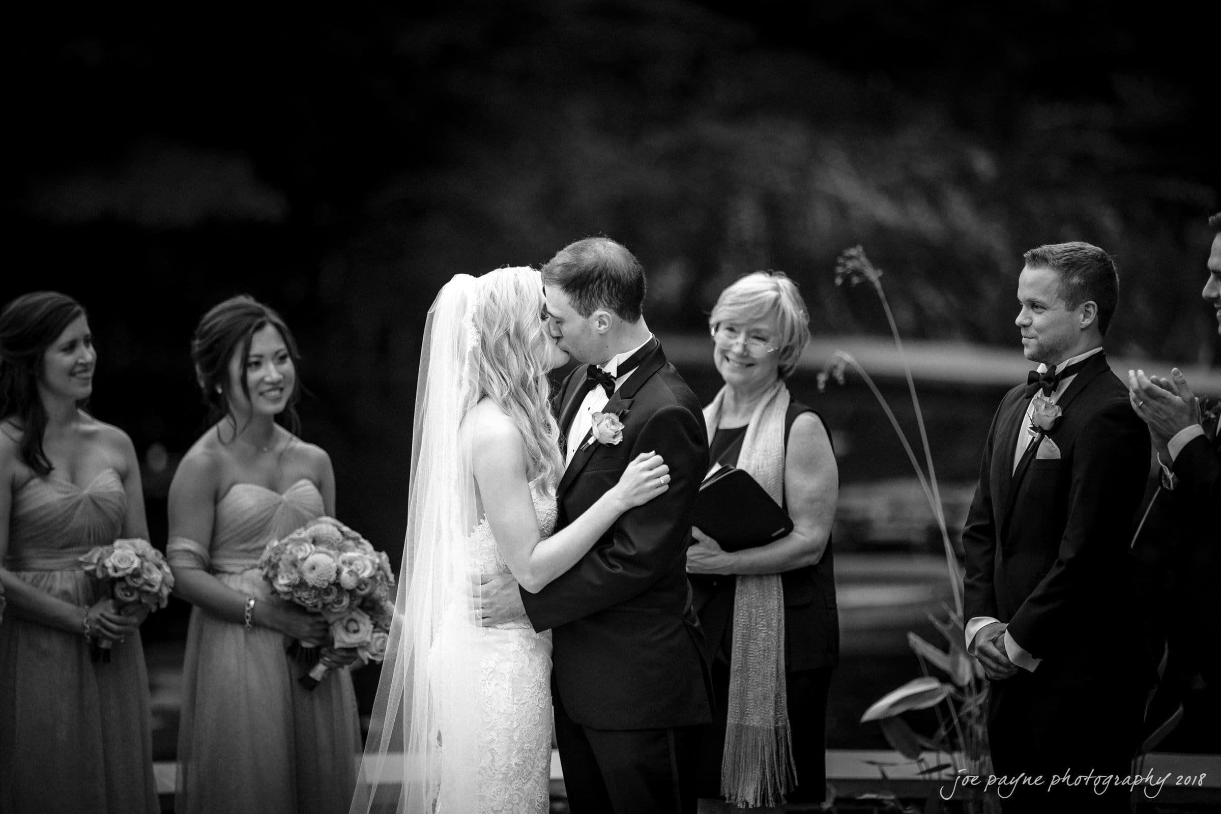 angle amphitheater at duke gardens wedding - katherine & andy A bride and groom share a kiss at their wedding ceremony. Bridesmaids holding bouquets stand on the left, and a groomsman and officiant are on the right. Everyone is dressed formally and the background is blurred.