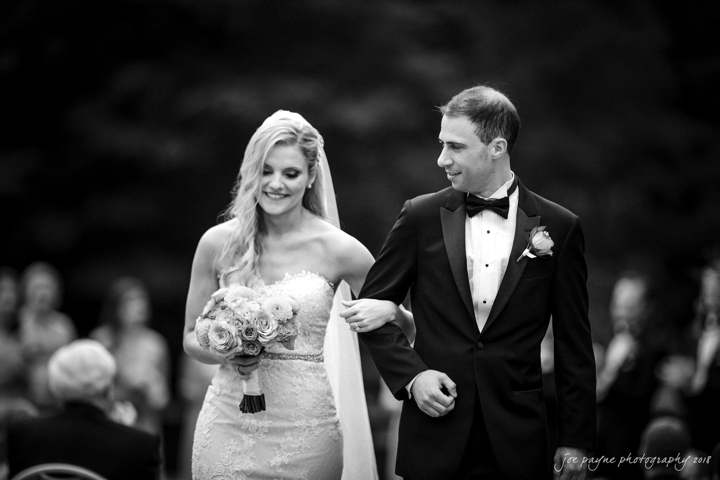 angle amphitheater at duke gardens wedding - katherine & andy A black and white photo of a smiling bride holding a bouquet and a groom in a tuxedo. They walk arm in arm down an aisle outdoors, with guests seated on either side. The bride has long hair and a strapless dress, while the groom wears a bowtie and boutonniere.