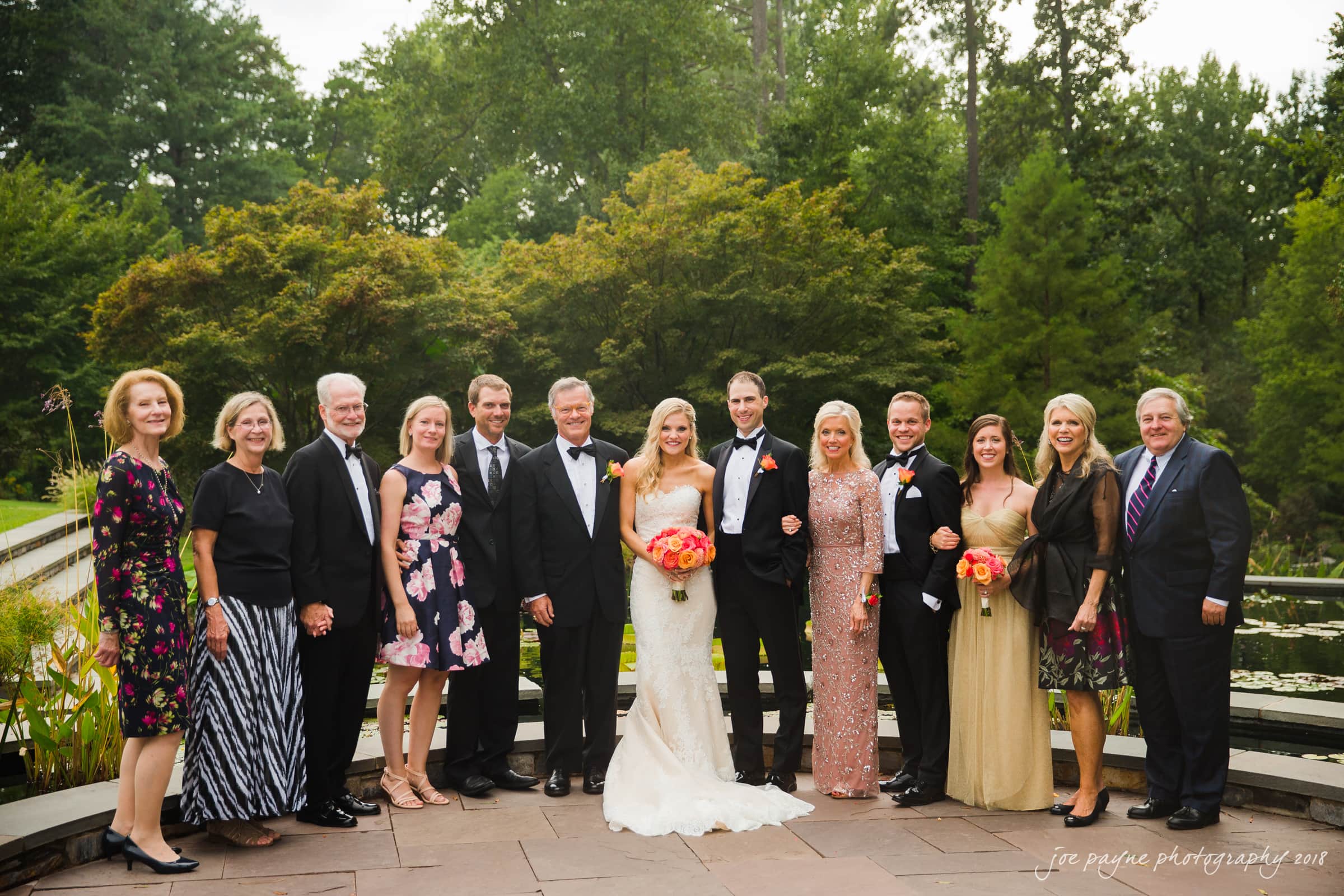 angle amphitheater at duke gardens wedding - katherine & andy A bride and groom stand together with family and friends in an outdoor setting. The group is dressed in formal attire, with the bride in a white gown and the groom in a black suit. They are posing for a photo in front of trees and greenery.