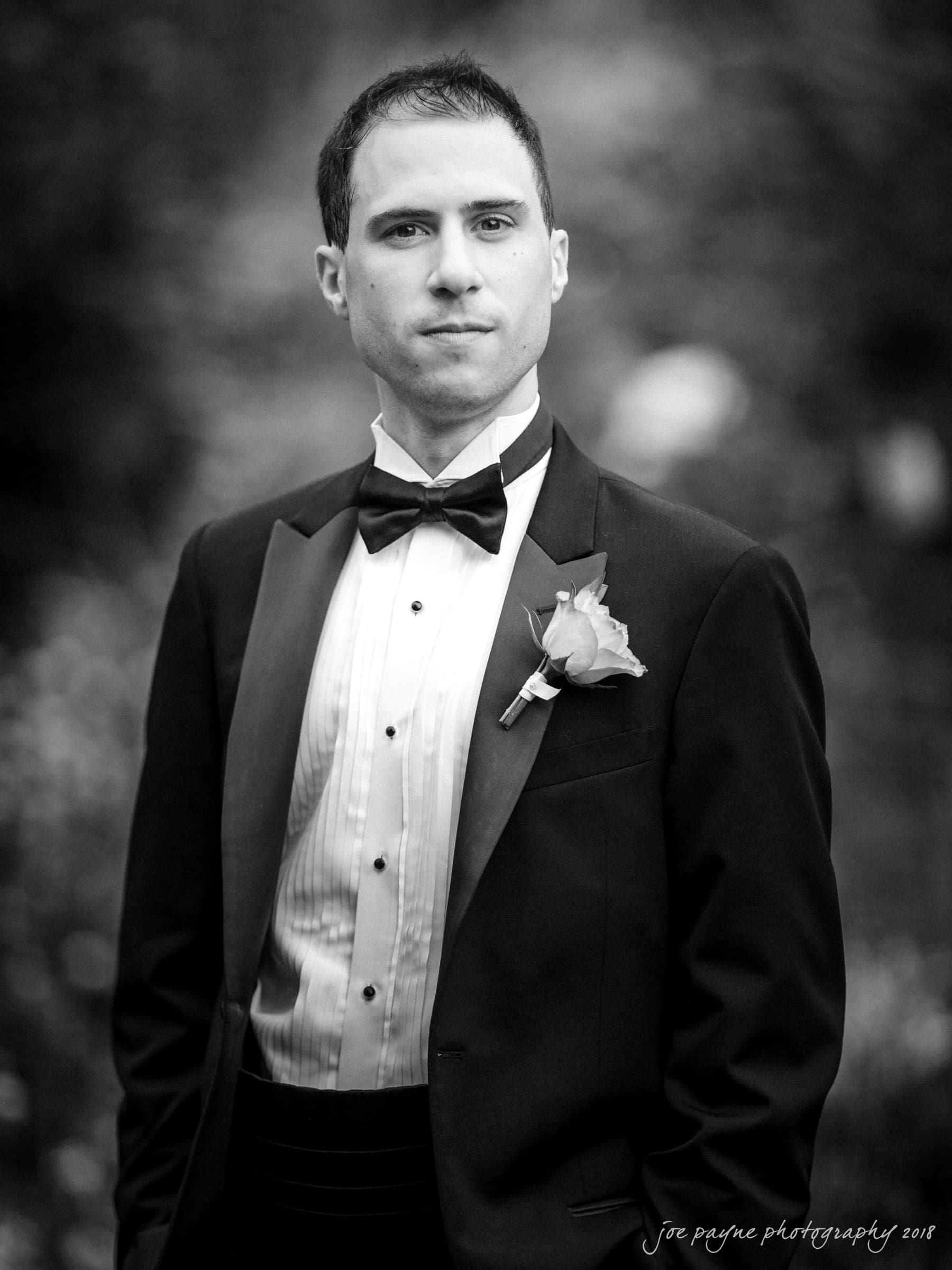 angle amphitheater at duke gardens wedding - katherine & andy A man in a tuxedo with a bow tie and a boutonniere stands outdoors. The black-and-white image captures him looking directly at the camera with a neutral expression. The background is blurred.
