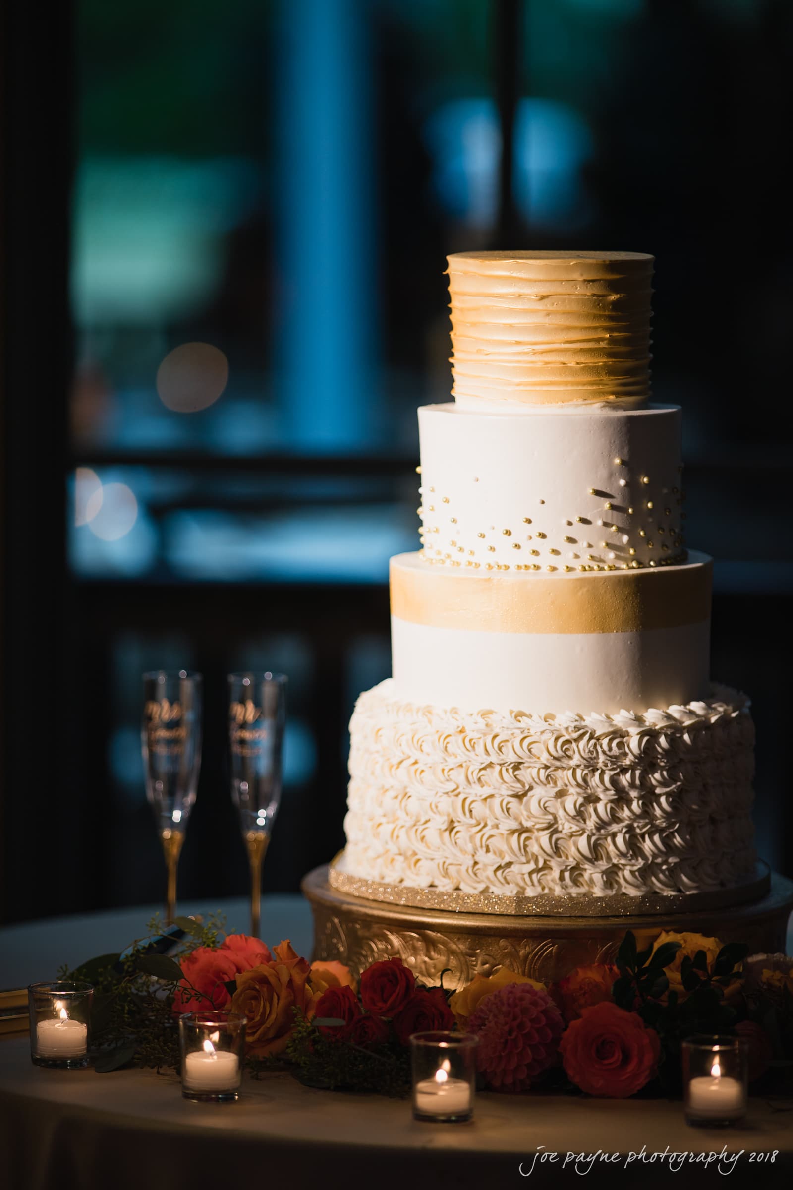 angle amphitheater at duke gardens wedding - katherine & andy A four-tier wedding cake with gold and white icing sits on a table adorned with flowers and candles. The top two tiers are gold, the third tier is decorated with gold dots, and the bottom tier features intricate white swirls.