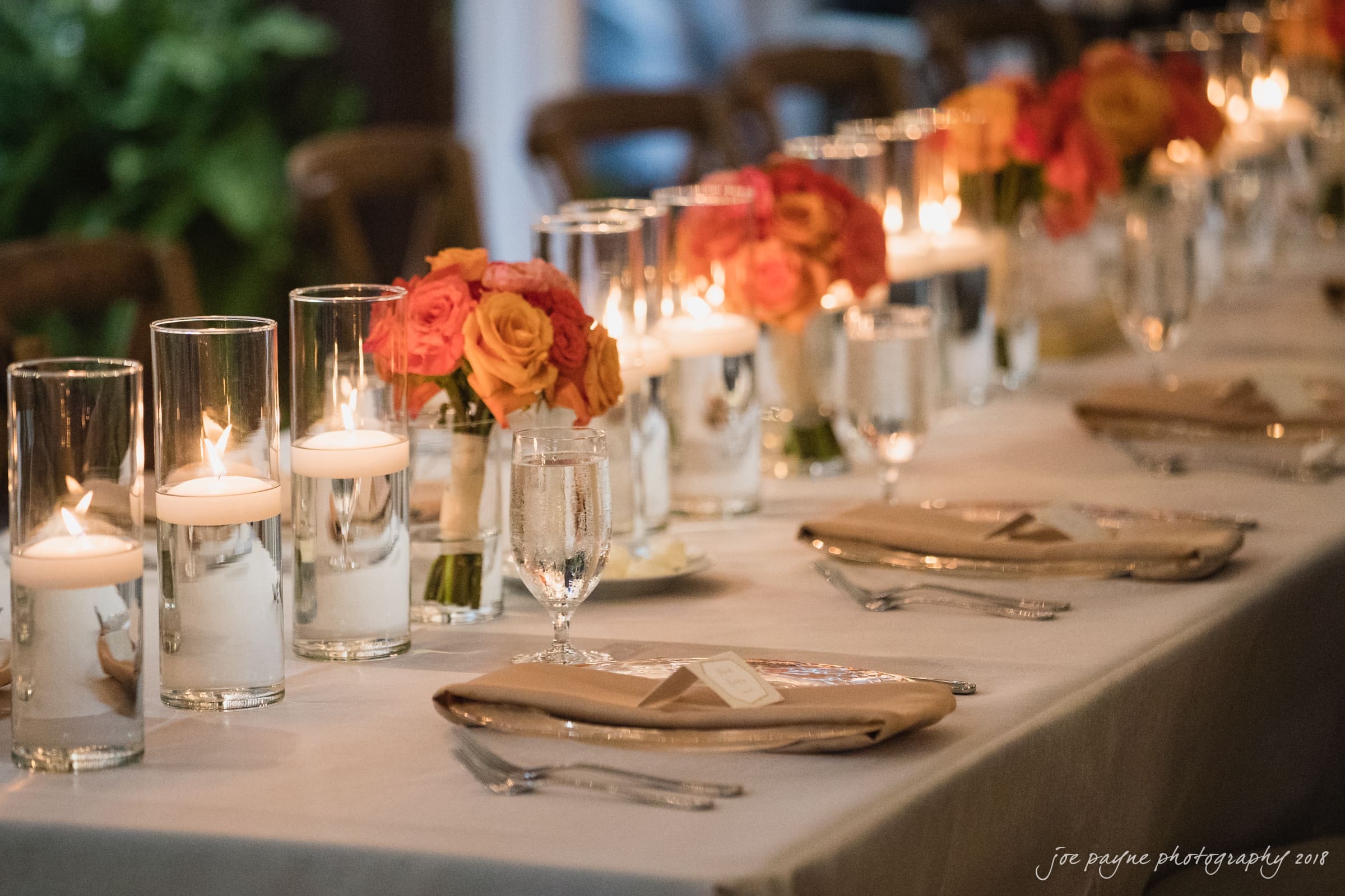 angle amphitheater at duke gardens wedding - katherine & andy Elegant table setting with a row of tall candles and orange roses in glass vases. Each place is set with beige napkins, cutlery, and glasses on a white tablecloth. The ambiance is warm and inviting, with soft candlelight.