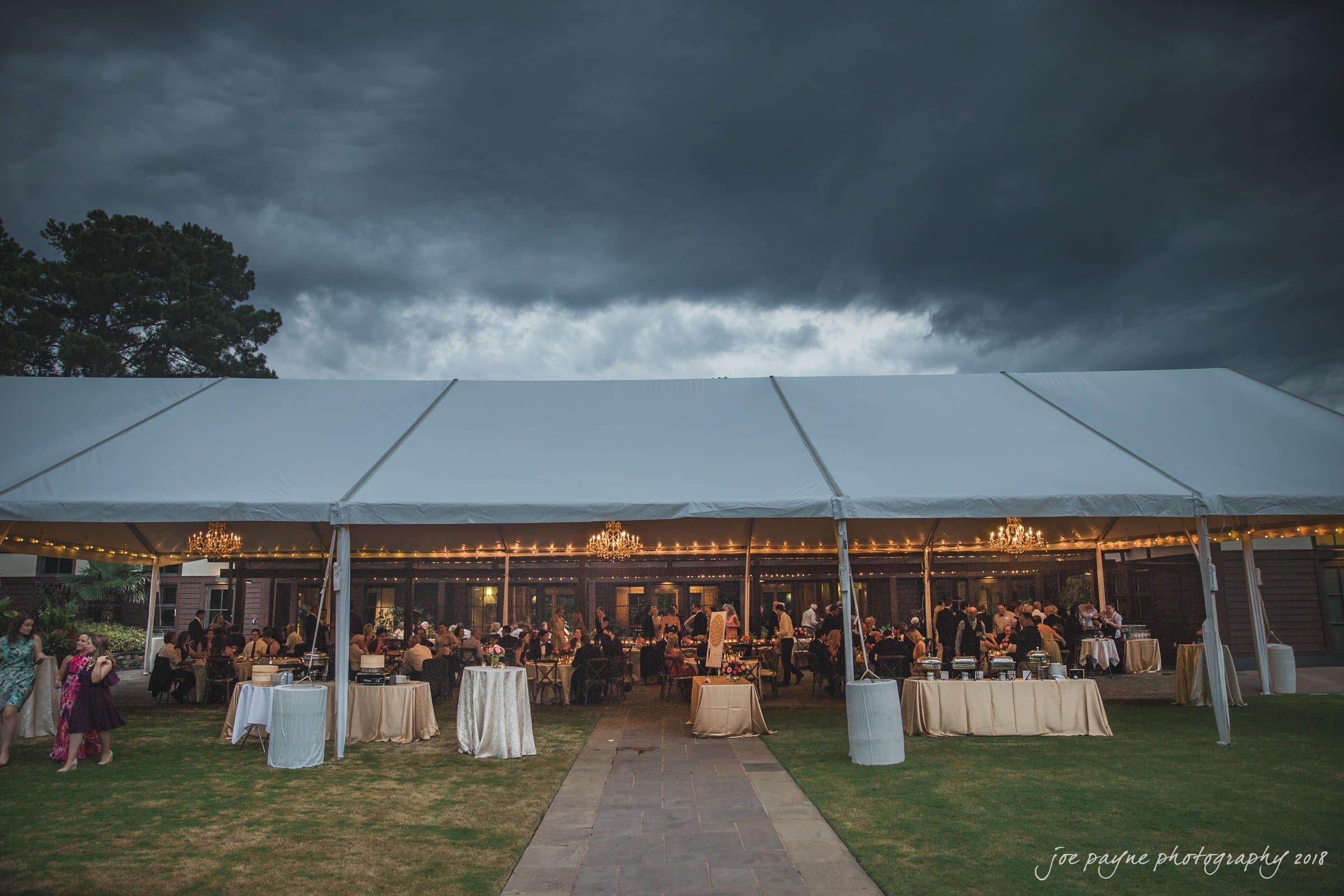 A wedding reception under a large white tent set up on grass. Guests dressed formally sit at tables decorated with white tablecloths. Chandeliers hang from the tent ceiling. The sky is overcast with dark, looming clouds.