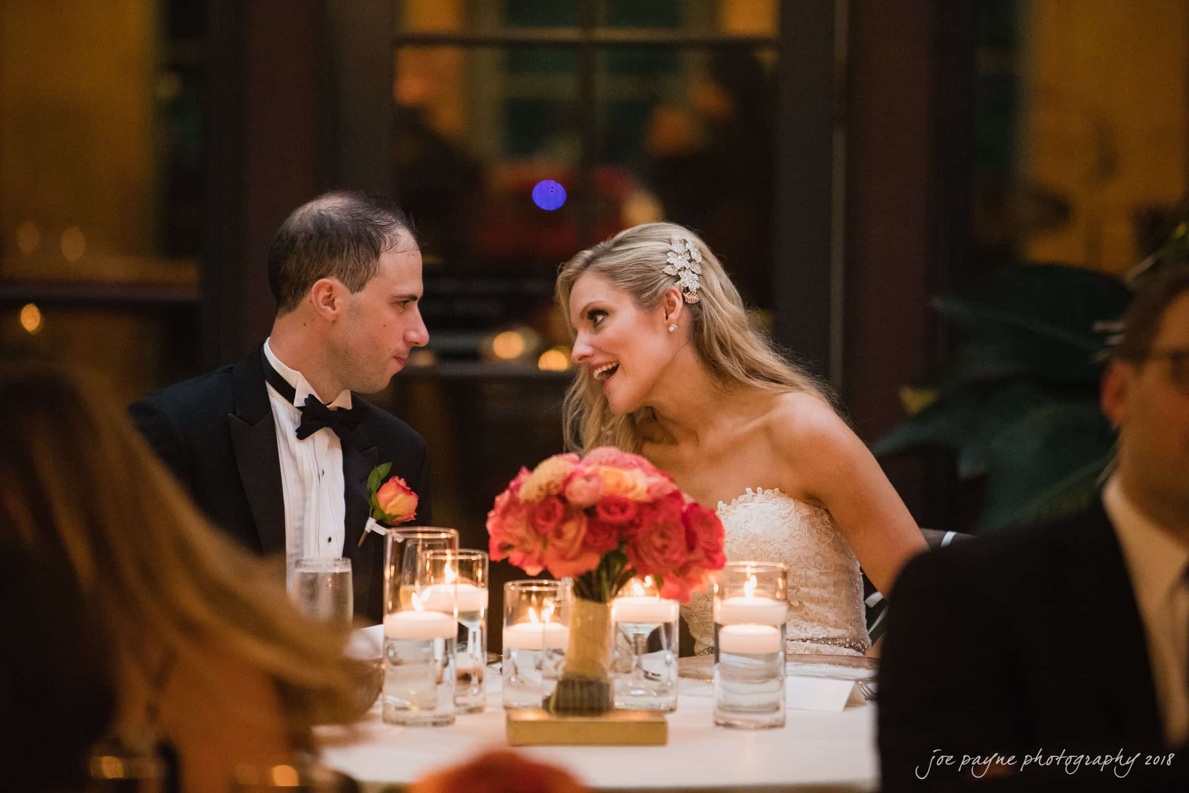angle amphitheater at duke gardens wedding - katherine & andy A bride and groom in formal attire sit at a table decorated with candles and flowers. The bride, wearing a white dress, smiles as she talks to the groom, who is in a black tuxedo with an orange boutonniere.
