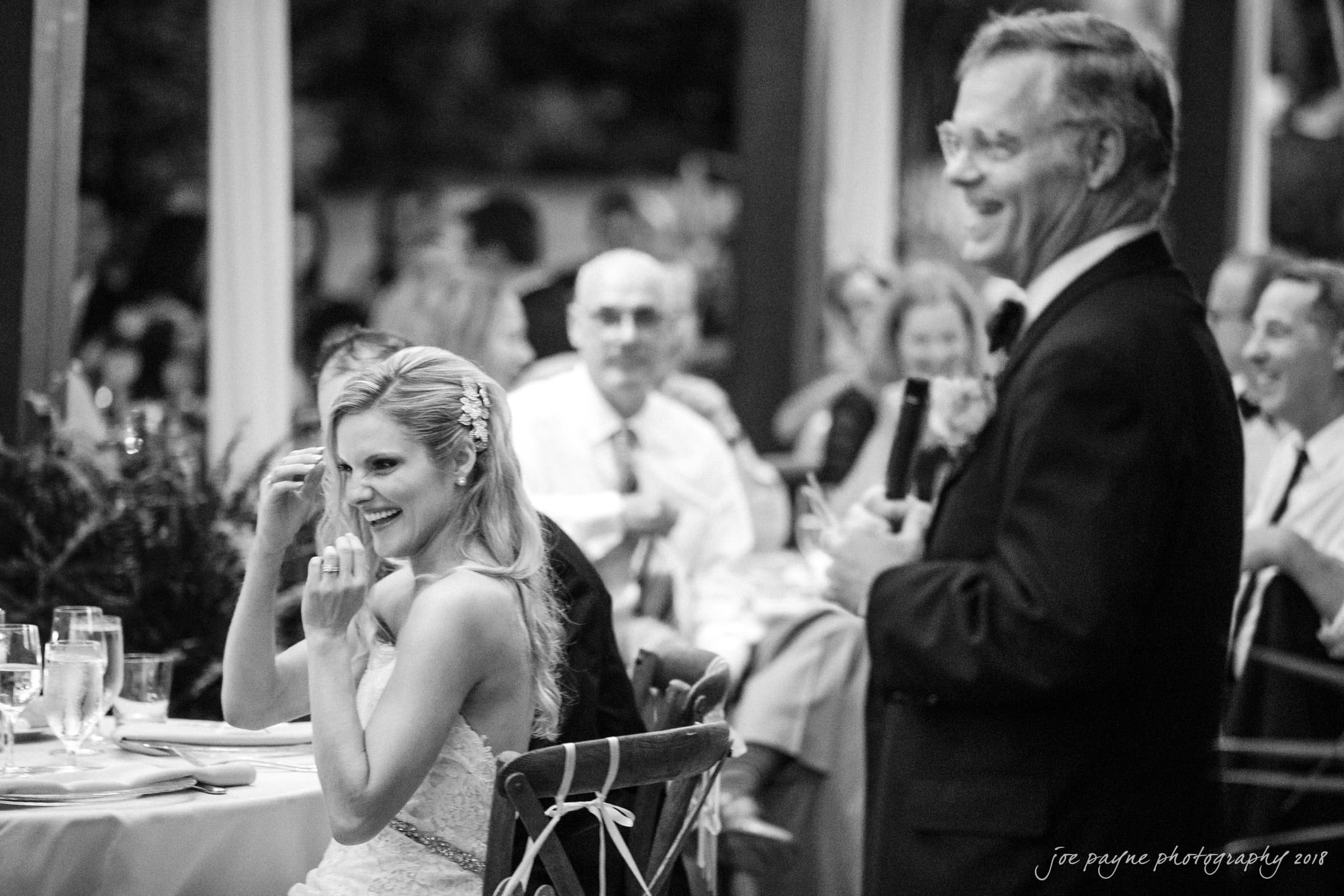 angle amphitheater at duke gardens wedding - katherine & andy A bride in a white dress laughs while seated at a wedding reception. An older man in a suit stands nearby, holding a microphone, smiling as he speaks. Guests are seated at tables in the background, enjoying the celebration.