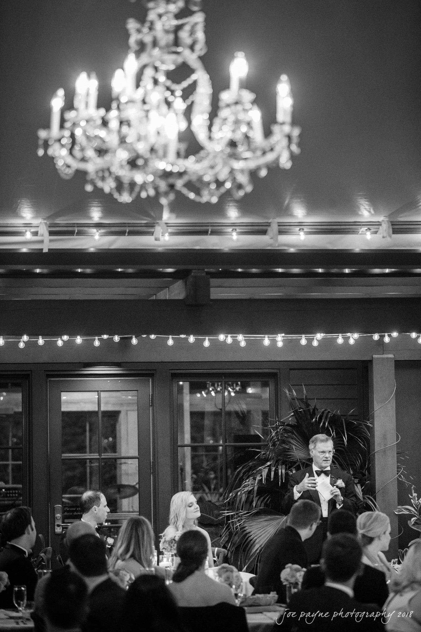 angle amphitheater at duke gardens wedding - katherine & andy A black and white image of a wedding reception. A chandelier hangs above a crowd of seated guests. One person stands, giving a speech. String lights decorate the area, and large windows are in the background.