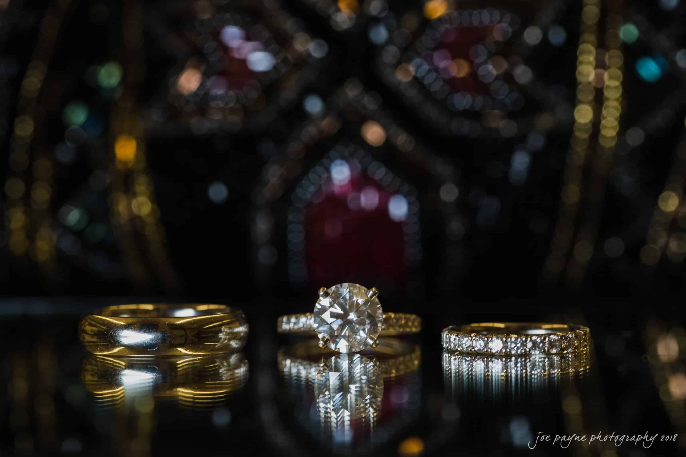 angle amphitheater at duke gardens wedding - katherine & andy Three rings are displayed against a dark, ornate background. The center ring features a large diamond, flanked by a gold band and a diamond-encrusted ring. The reflective surface emphasizes their elegance.