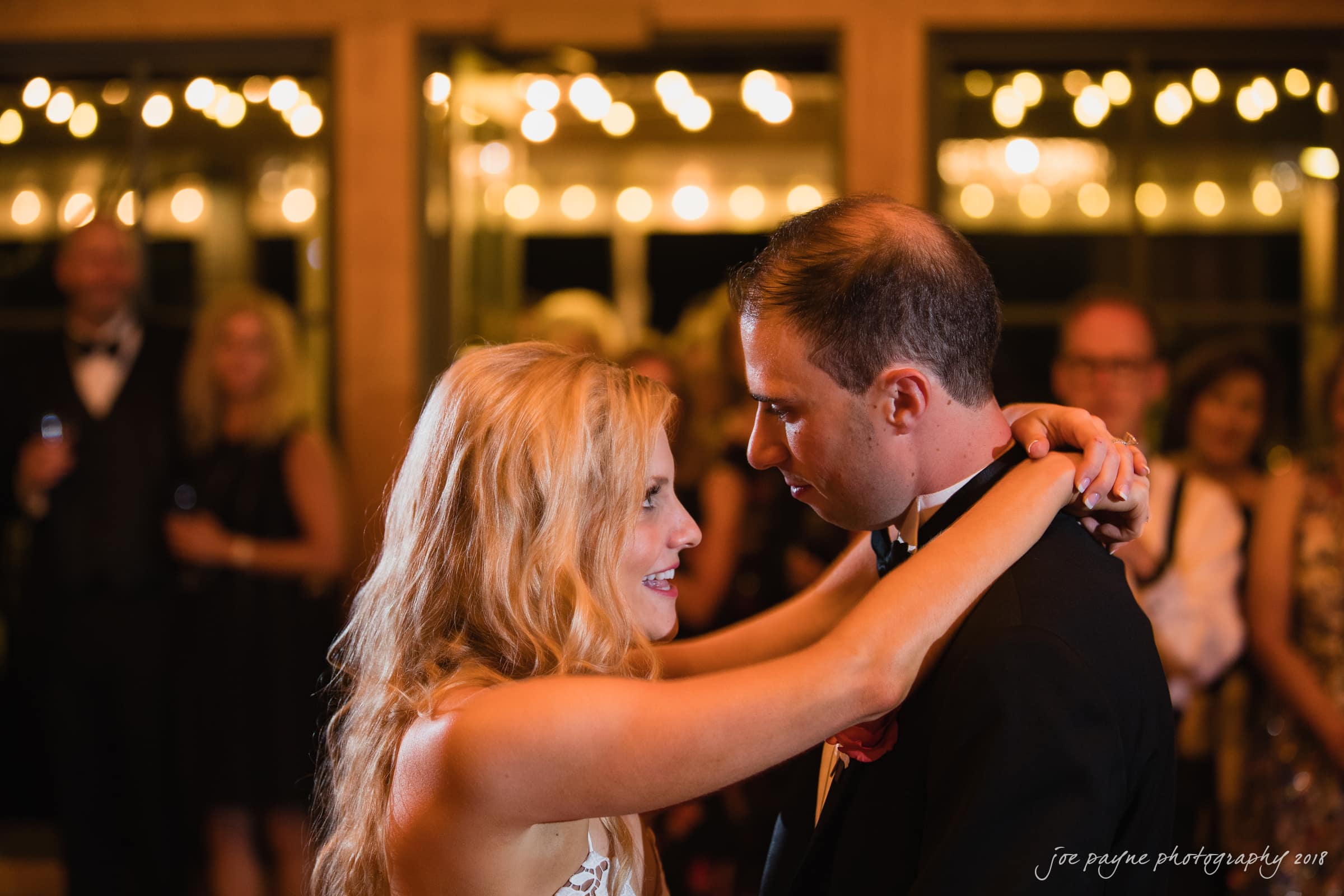 angle amphitheater at duke gardens wedding - katherine & andy A bride and groom dance closely, gazing into each others eyes. The background is softly blurred, filled with warm lights and elegantly dressed guests, creating a romantic atmosphere.