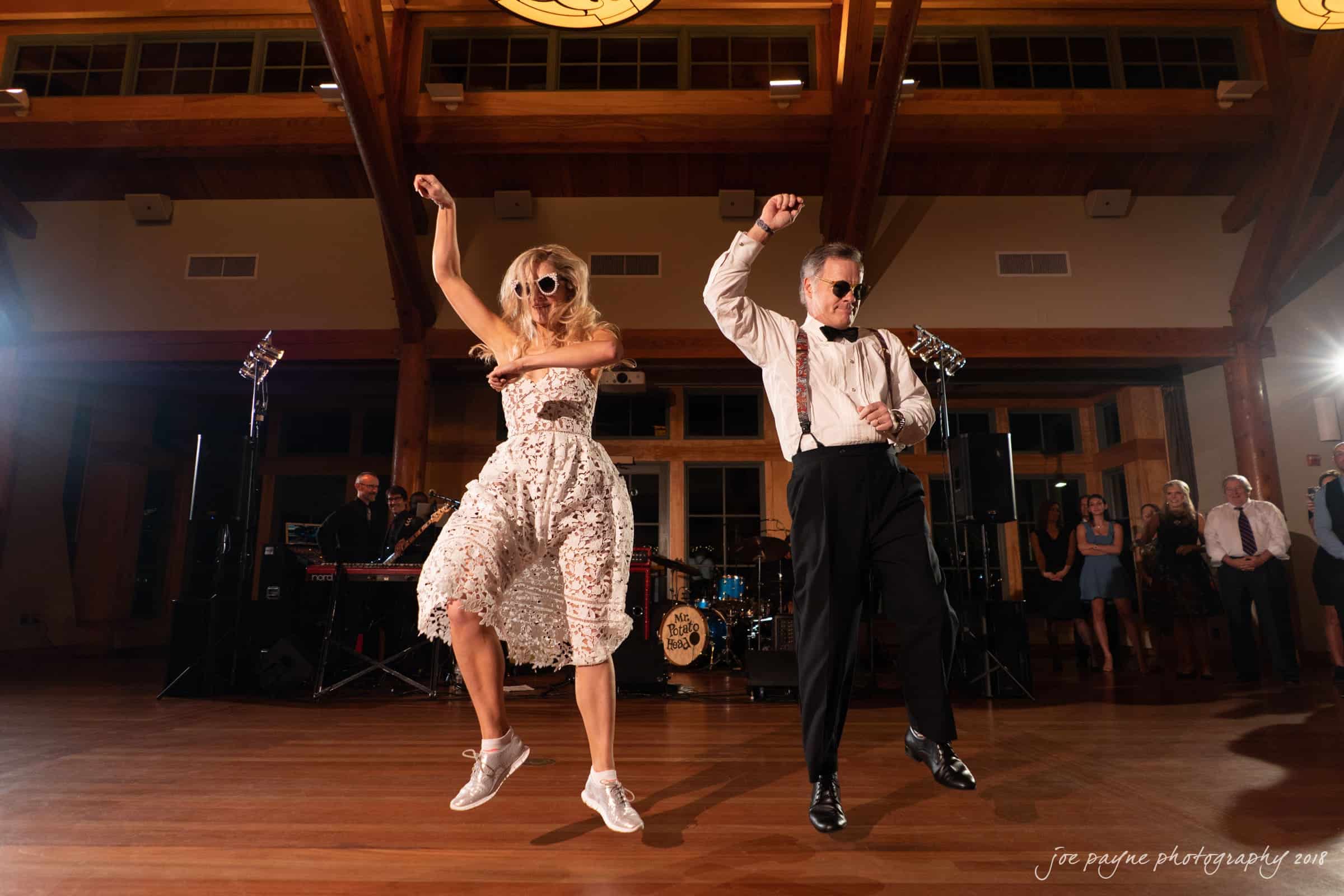 angle amphitheater at duke gardens wedding - katherine & andy A man and a woman energetically dancing on a wooden floor in a spacious hall, illuminated by warm lighting. The man wears a white shirt with suspenders, while the woman is in a white dress. A band setup is visible in the background.