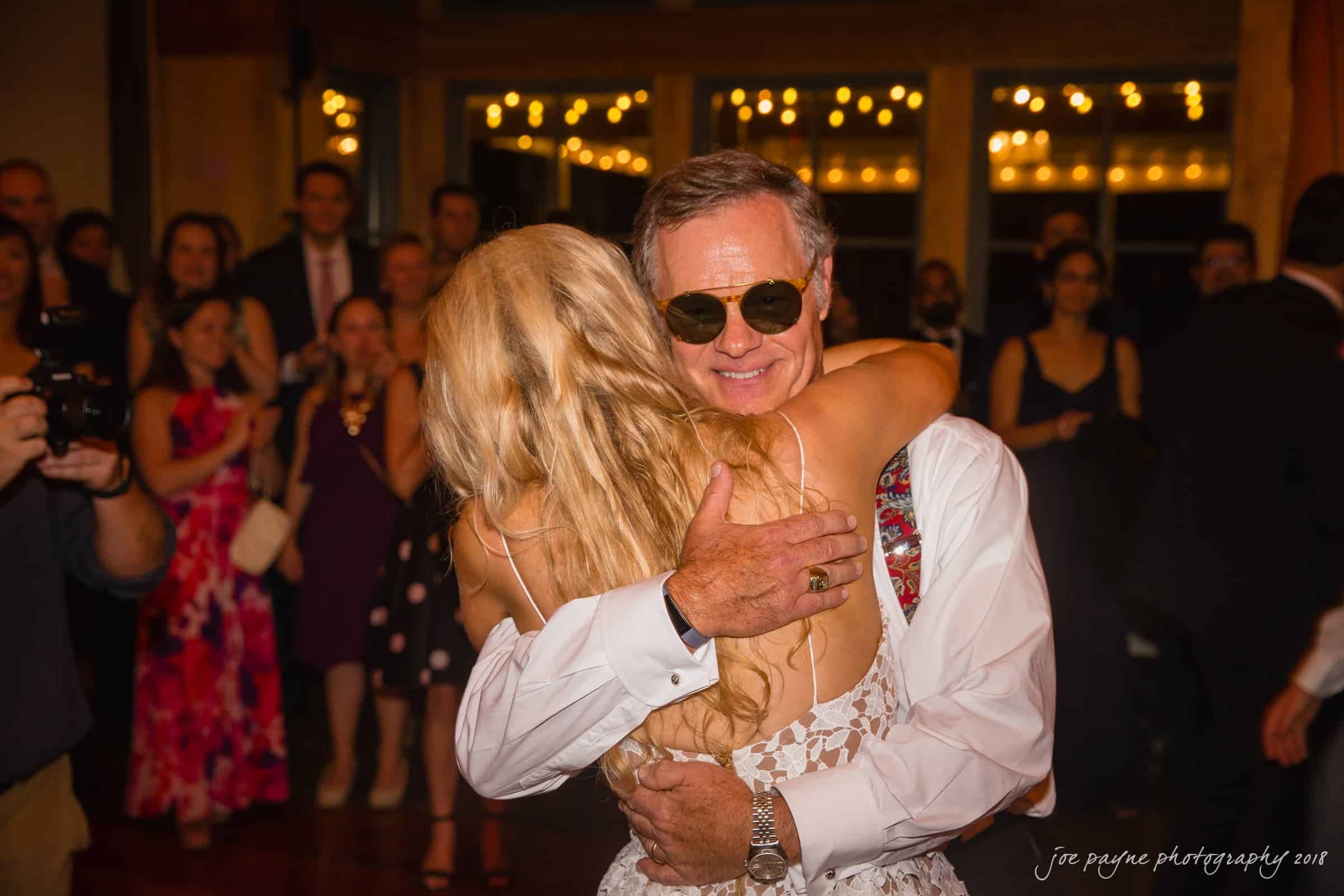 angle amphitheater at duke gardens wedding - katherine & andy A joyful moment at a wedding reception as a man in sunglasses embraces a woman with long blonde hair. Guests are gathered in the background, some smiling, with warm lighting and festive decorations.