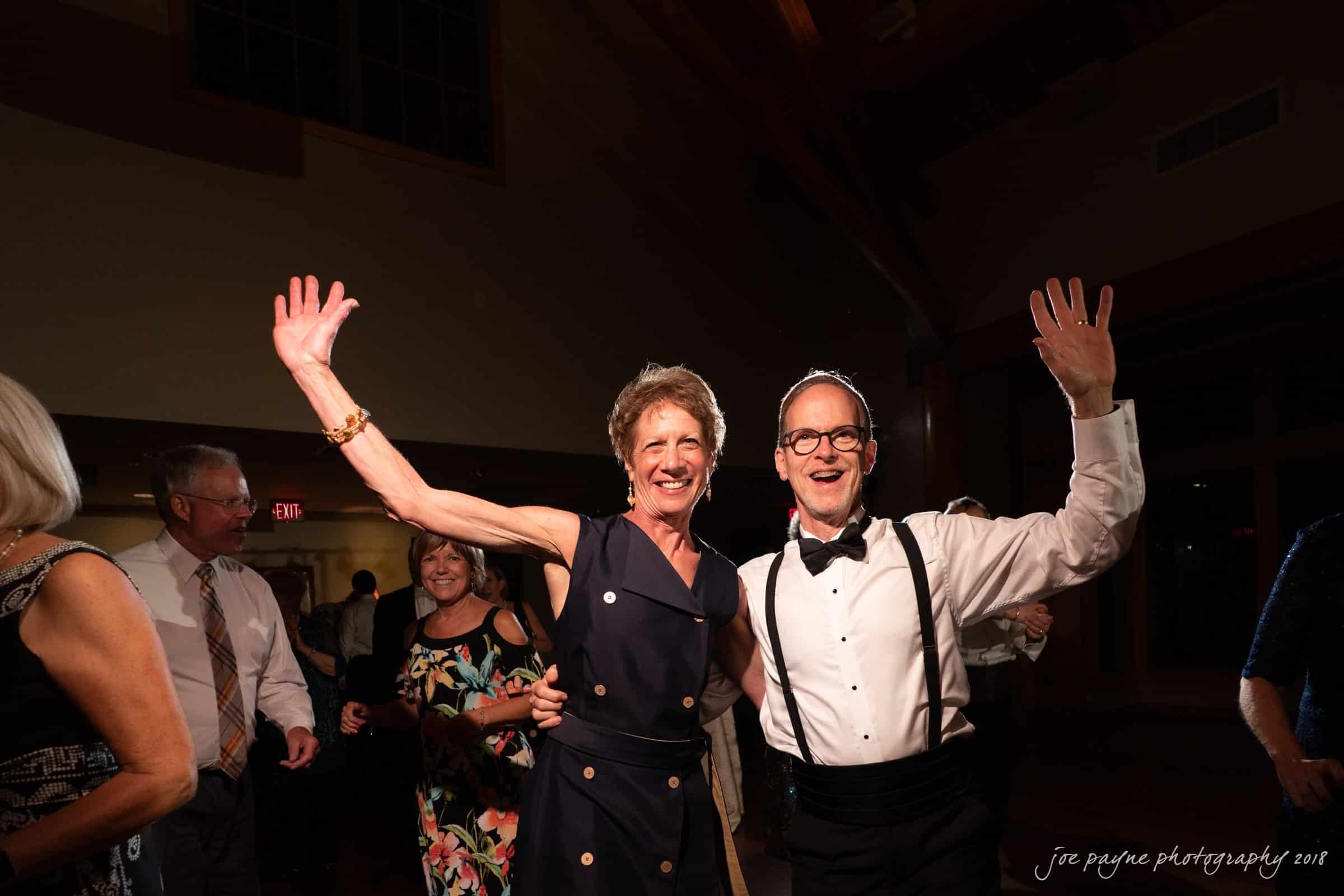 angle amphitheater at duke gardens wedding - katherine & andy A couple in formal attire poses happily at a lively indoor event. The woman in a navy dress and the man in a white shirt with suspenders both raise their arms, smiling broadly. Other guests are visible in the background under warm lighting.