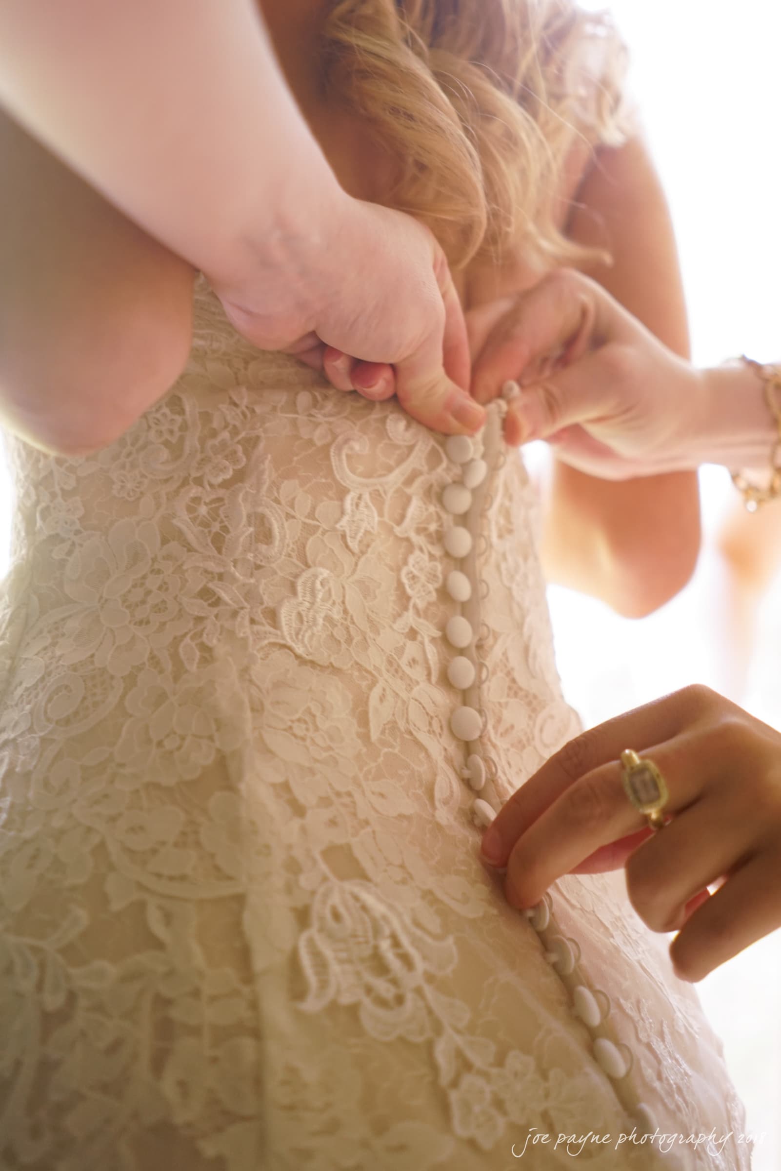 angle amphitheater at duke gardens wedding - katherine & andy Close-up of three hands fastening buttons on the back of a white lace wedding dress. The brides blonde curls are visible, and light gently illuminates the scene, creating a warm and intimate atmosphere.