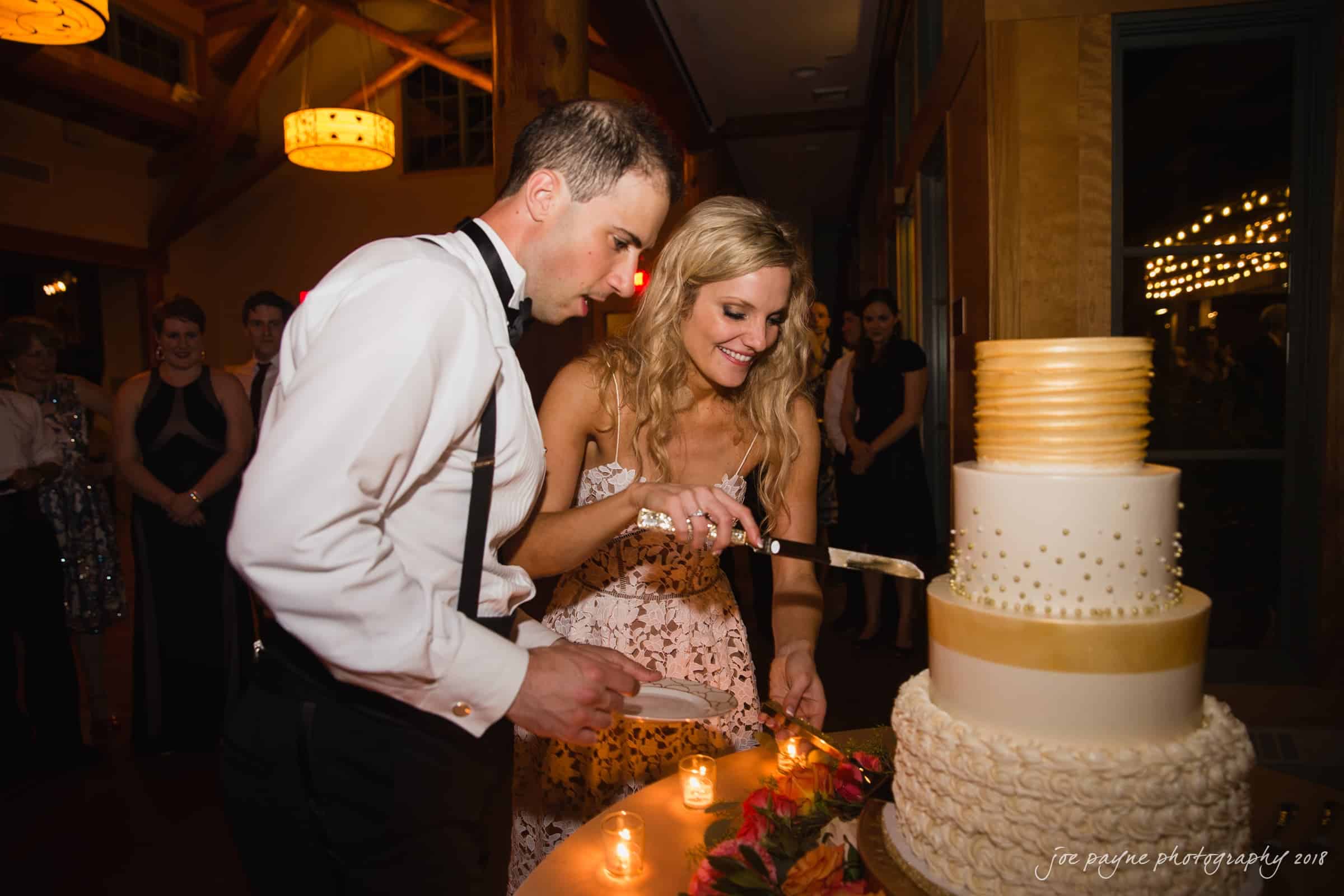 angle amphitheater at duke gardens wedding - katherine & andy A bride and groom are cutting a three-tiered wedding cake with gold accents. They are surrounded by guests in a warmly lit venue. Candles and autumn foliage decorate the table. Both are smiling and wearing formal attire.
