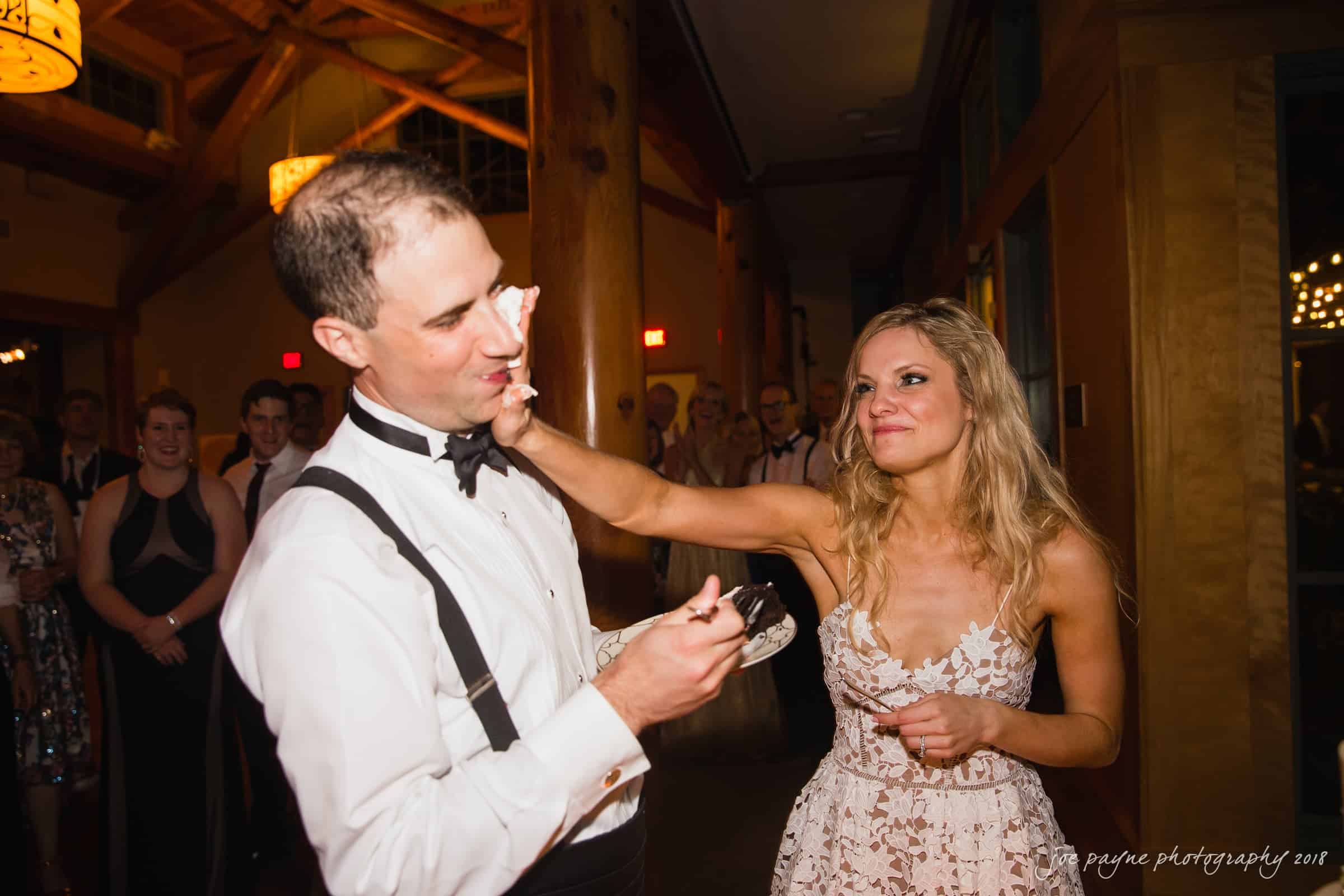angle amphitheater at duke gardens wedding - katherine & andy A woman playfully smears cake on a mans face during a wedding reception. The man is dressed in a tuxedo, and the woman is in a patterned dress. They are surrounded by guests in a warmly lit venue.