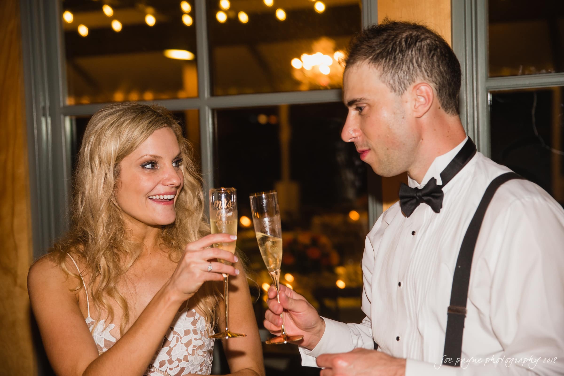 angle amphitheater at duke gardens wedding - katherine & andy A woman in a white lace dress and a man in a white shirt with suspenders toast with champagne glasses. They are indoors, and warm lighting from behind creates a festive atmosphere.