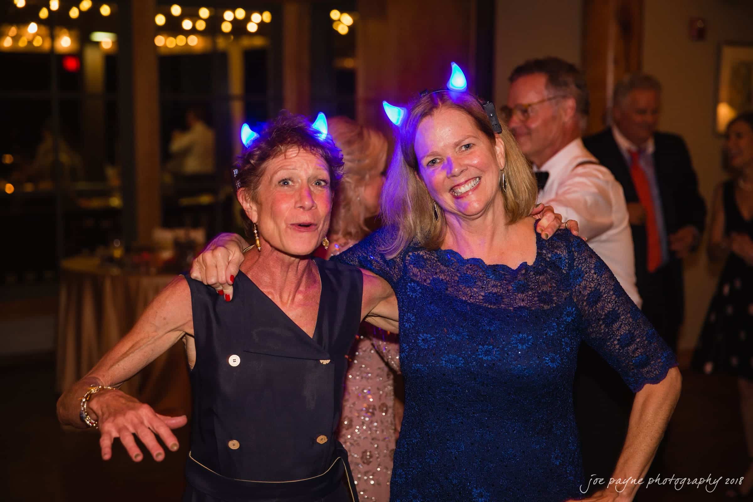 angle amphitheater at duke gardens wedding - katherine & andy Two women, one in a black dress and the other in a blue lace dress, both wearing glowing blue devil horn headbands, smiling and posing arm-in-arm at a party. Other guests are seen in the dimly lit background.