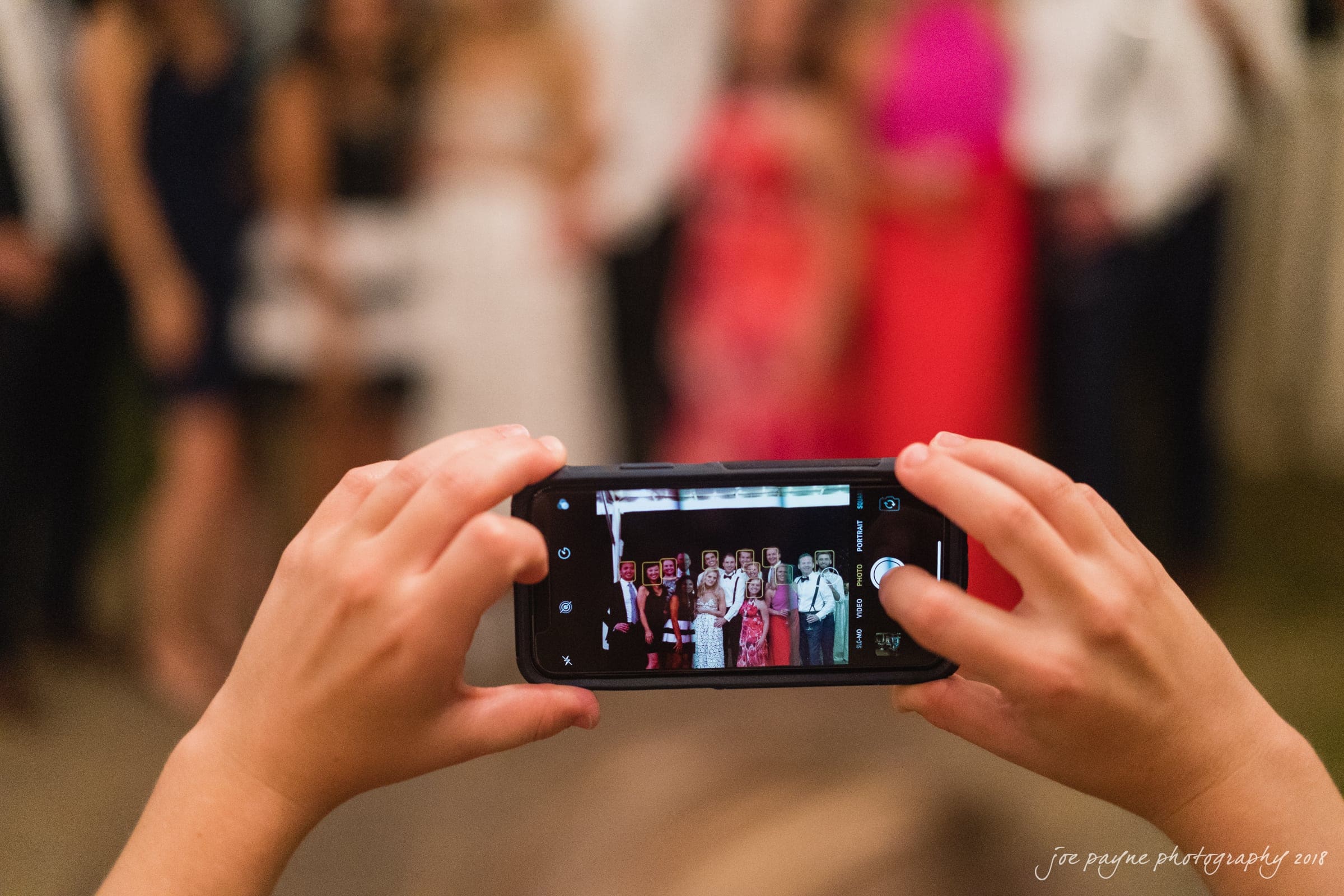 angle amphitheater at duke gardens wedding - katherine & andy A person holds a smartphone, capturing a group photo of several people dressed formally. The image on the phone is in focus, showing smiles and various colored outfits. The background is blurred.