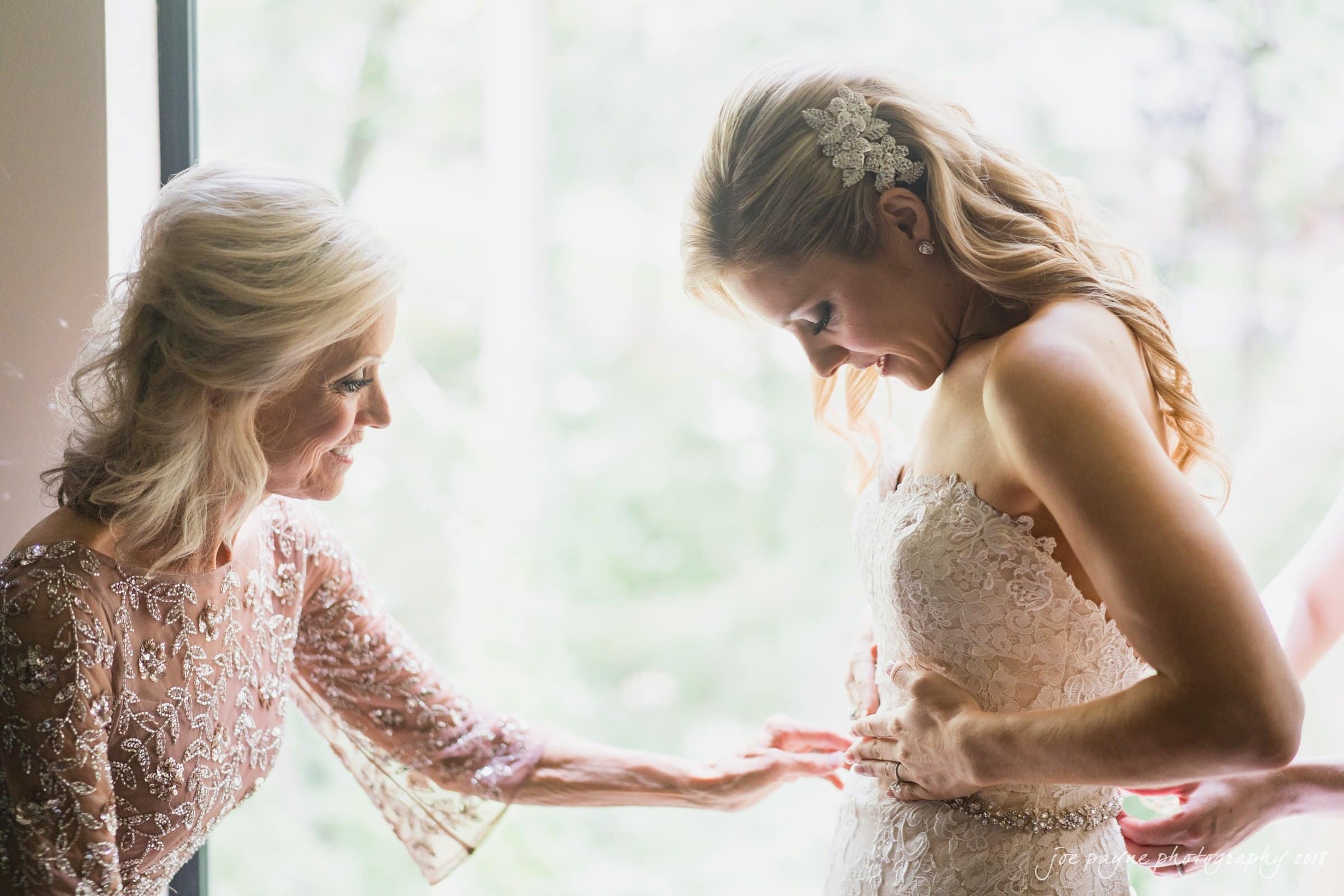 angle amphitheater at duke gardens wedding - katherine & andy A bride in a lace wedding dress is being helped by an older woman in an elegant lace dress. They stand near a window with natural light highlighting their expressions of focus and care.