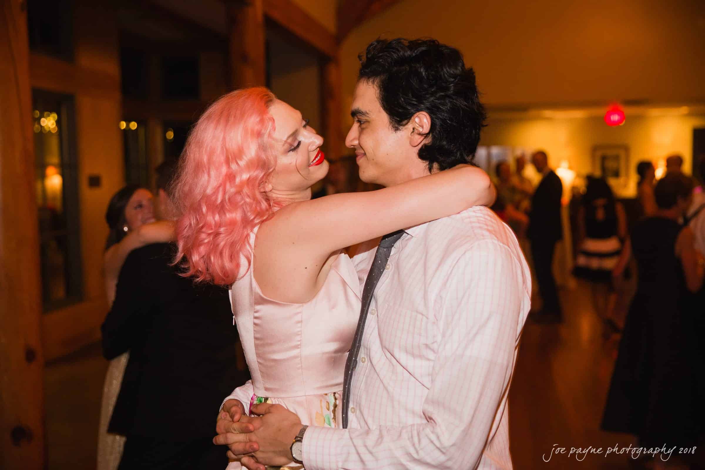 angle amphitheater at duke gardens wedding - katherine & andy A couple embraces and smiles at each other on a dimly lit dance floor. The woman has pink hair and wears a light pink dress, while the man is in a dress shirt and tie. Others can be seen dancing in the background.