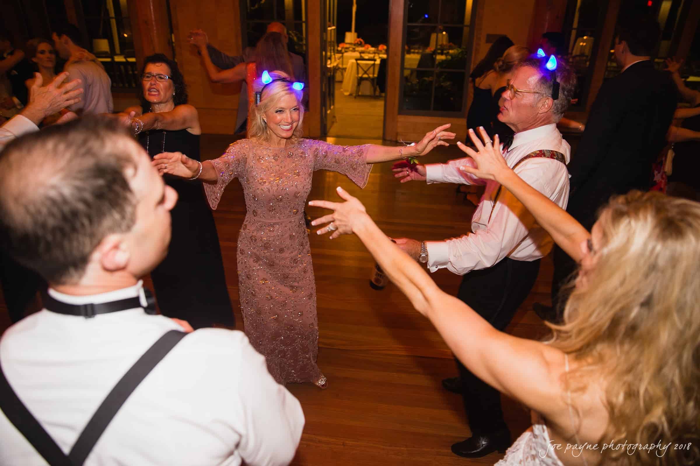 angle amphitheater at duke gardens wedding - katherine & andy A group of people dancing energetically on a wooden floor. A woman in a lace dress is at the center, with several others wearing formal attire surrounding her. Some are wearing light-up headbands, creating a festive atmosphere.
