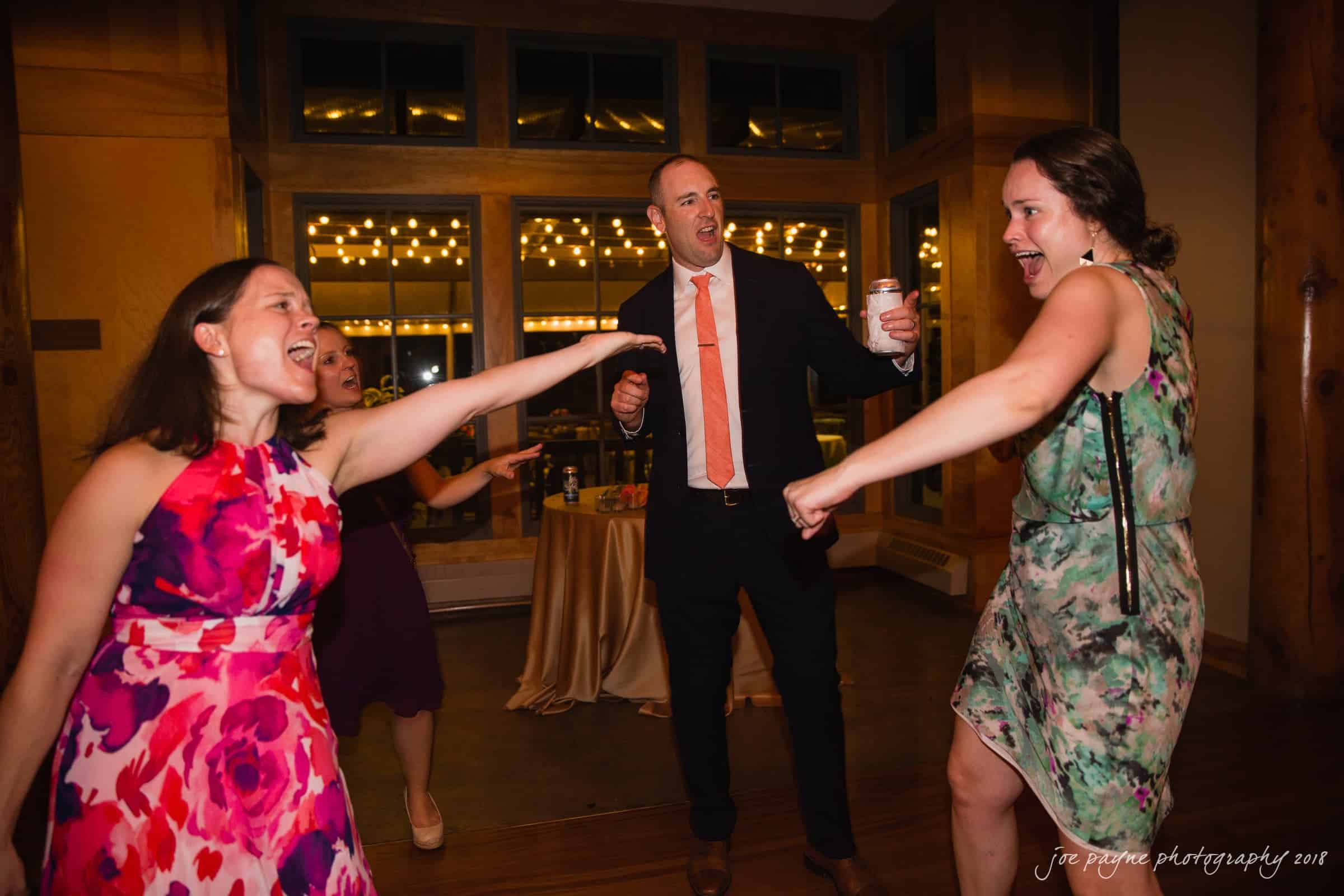 angle amphitheater at duke gardens wedding - katherine & andy Three people in colorful dresses and a suit dance energetically at a party. One woman points forward, and the man holds a drink. Warm lighting and string lights create a festive atmosphere in the background.