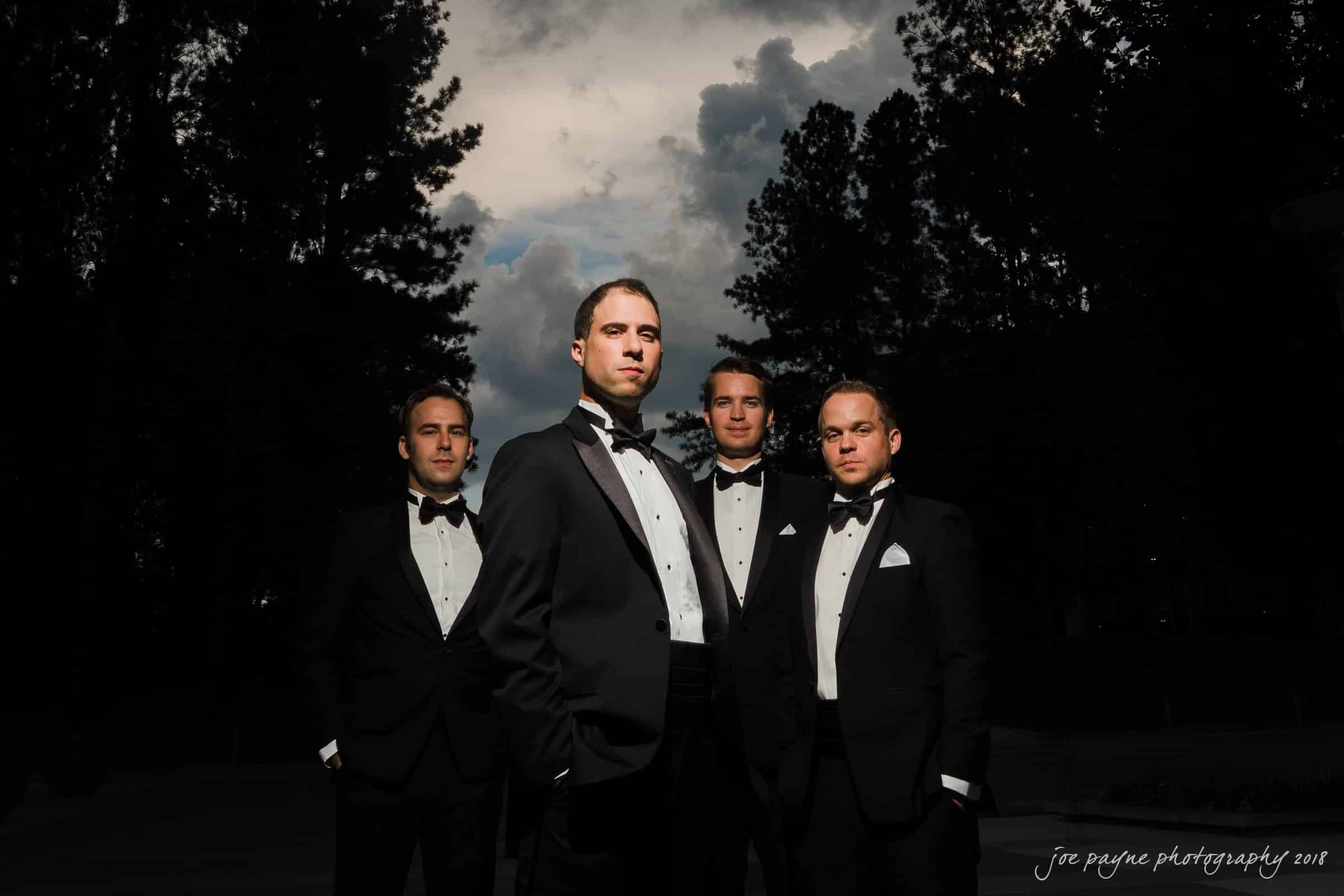 angle amphitheater at duke gardens wedding - katherine & andy Four men wearing tuxedos stand confidently outdoors against a backdrop of tall trees and a dramatic cloudy sky. The lighting highlights the central figure, creating a formal and striking composition.