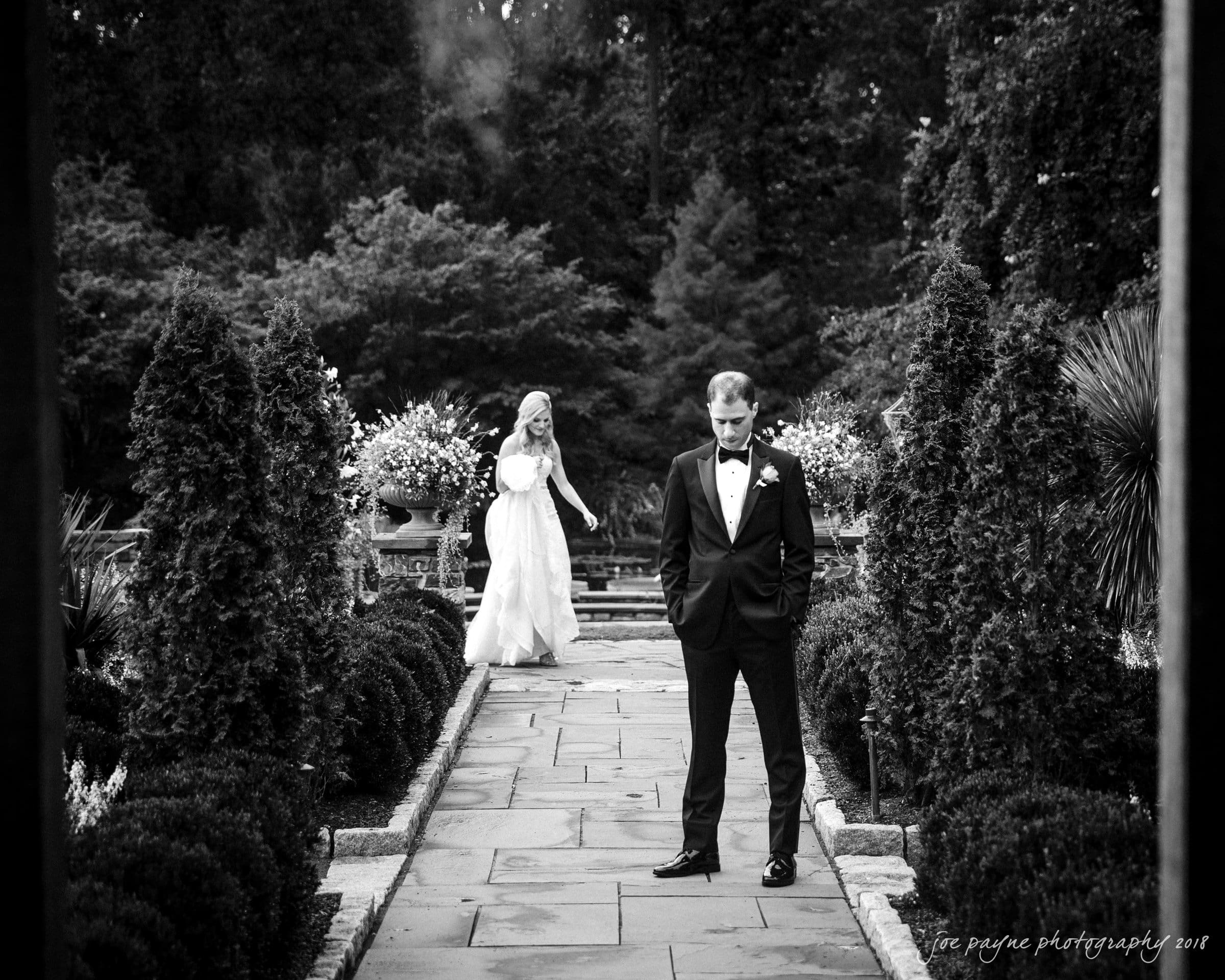 angle amphitheater at duke gardens wedding - katherine & andy A bride playfully approaches a groom standing on a garden pathway, obscured by a hedge. The groom looks down, unaware of her presence, creating a surprise moment amidst lush greenery. The scene is captured in black and white.