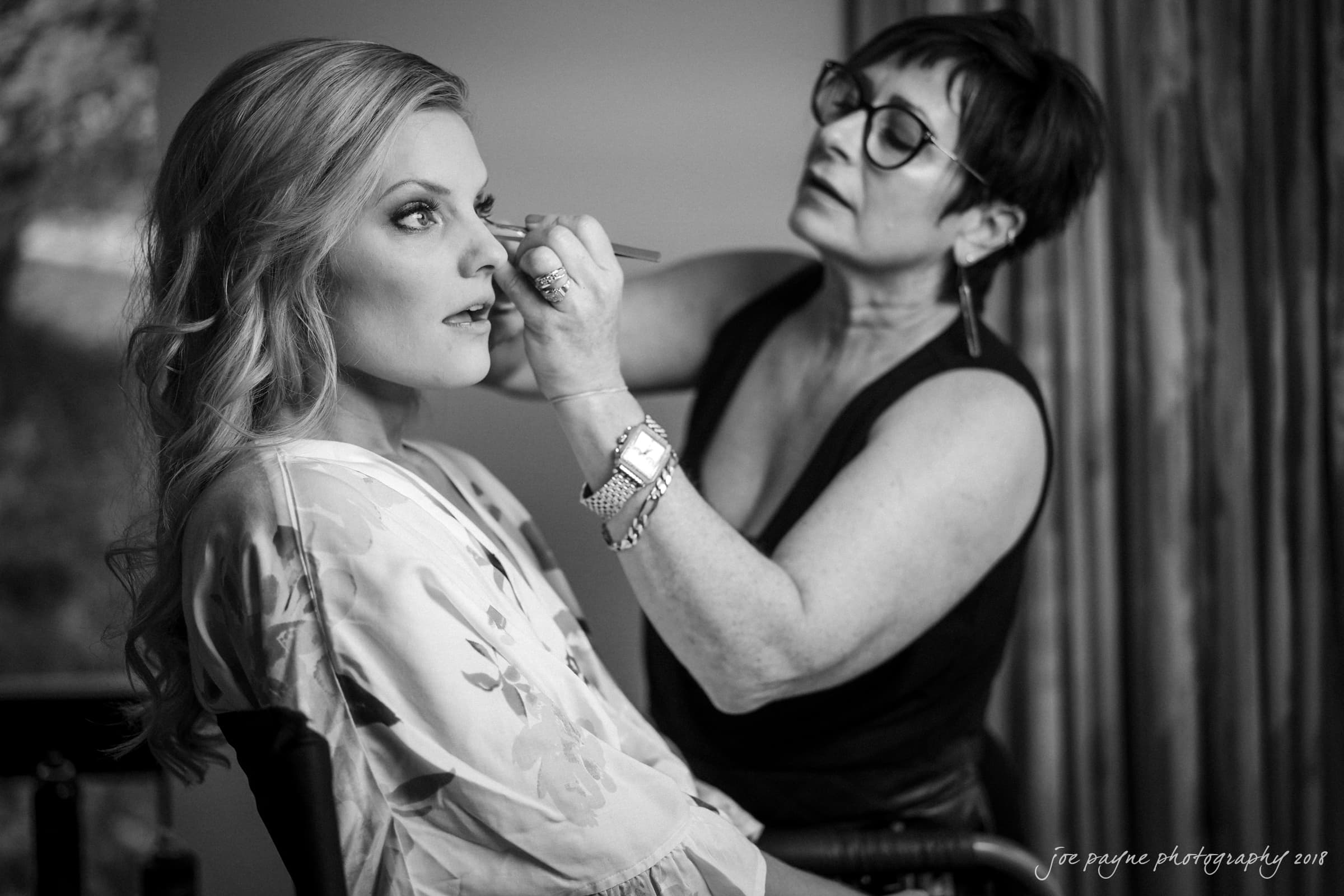 angle amphitheater at duke gardens wedding - katherine & andy A black and white photo of a woman having her makeup done by a makeup artist. The woman is sitting in a chair wearing a floral robe, while the artist, standing beside her, applies makeup with a brush.