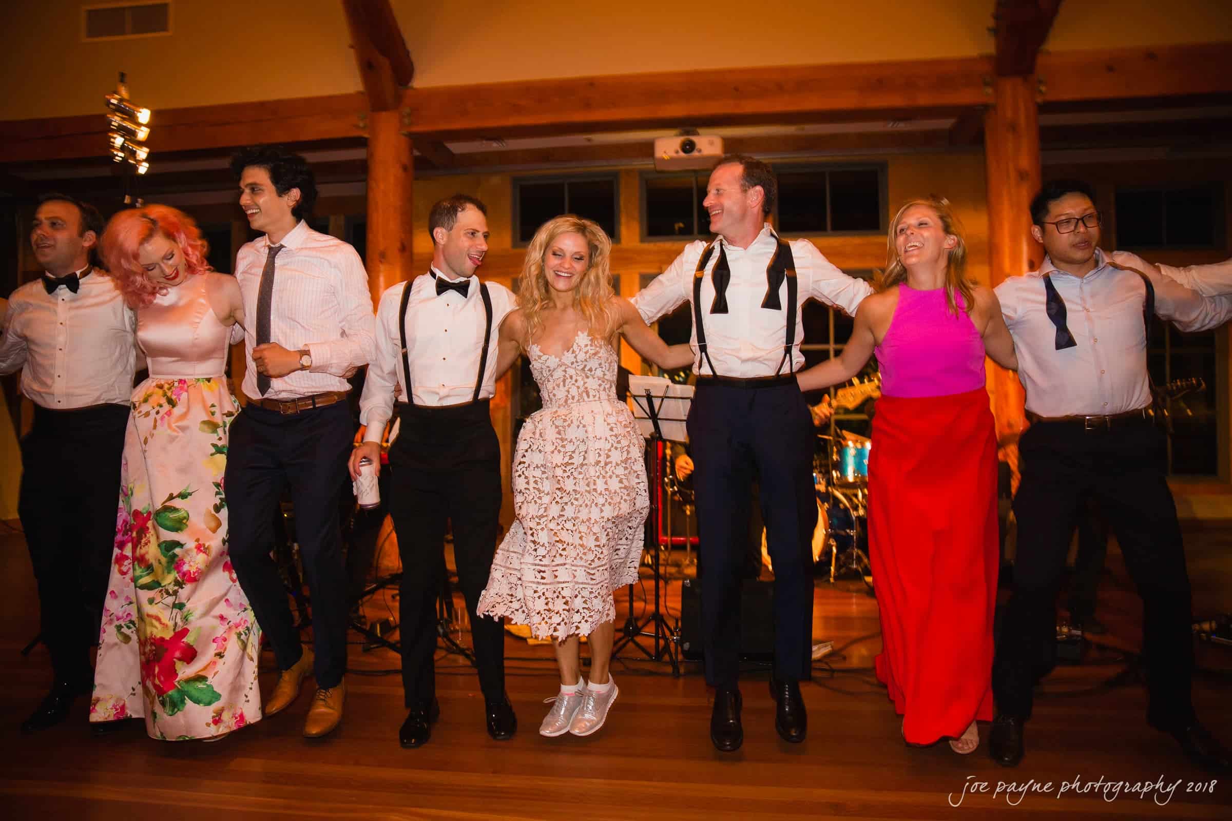 angle amphitheater at duke gardens wedding - katherine & andy A group of people in formal attire stand in a line with their arms around each other, smiling and looking joyful. They are in a warmly lit room with wooden beams. A live band setup is visible in the background.