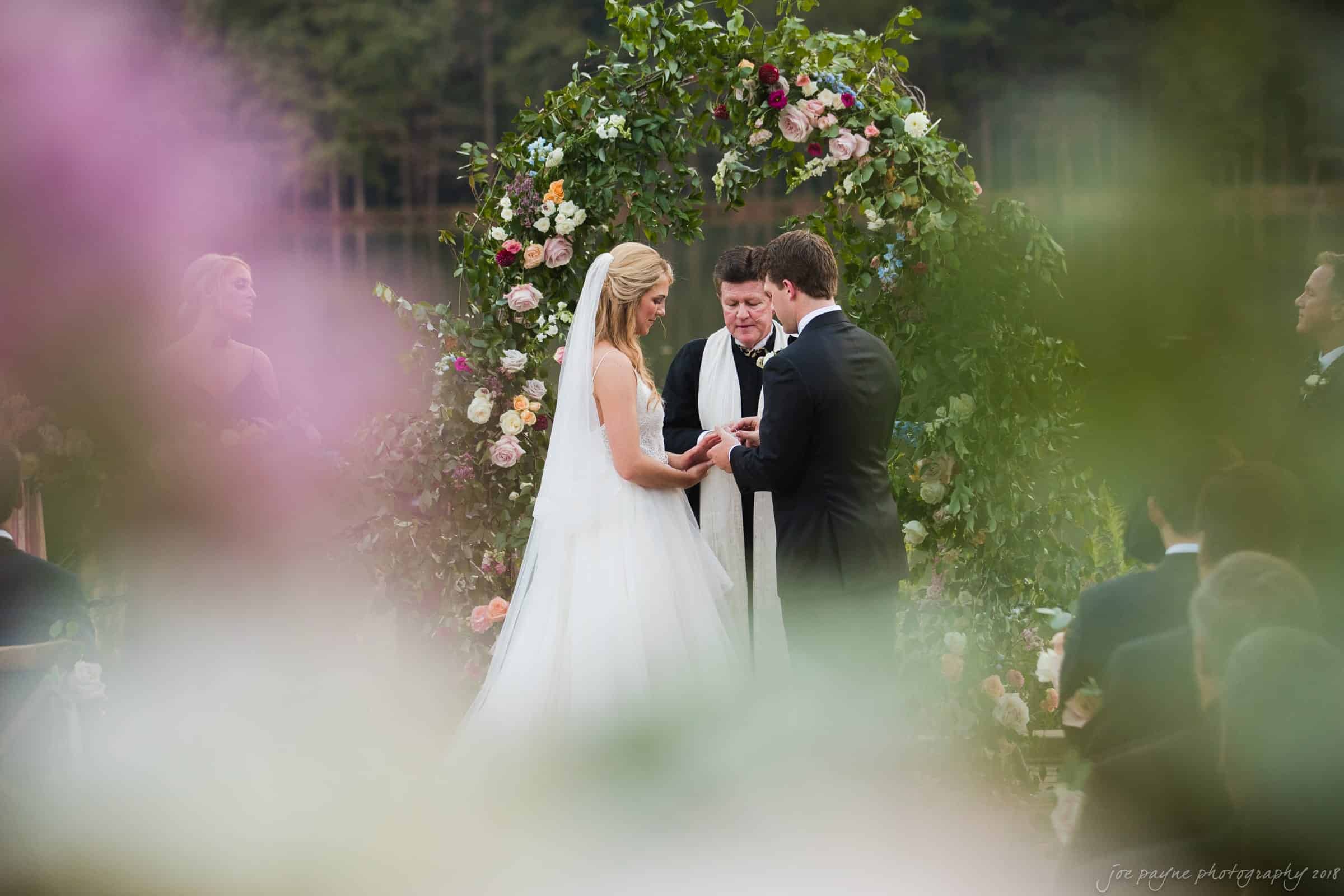 A bride and groom stand facing each other, exchanging vows in an outdoor ceremony. They are under a floral arch, with a lush garden backdrop. Guests are seated nearby, partially obscured by flowers in the foreground.