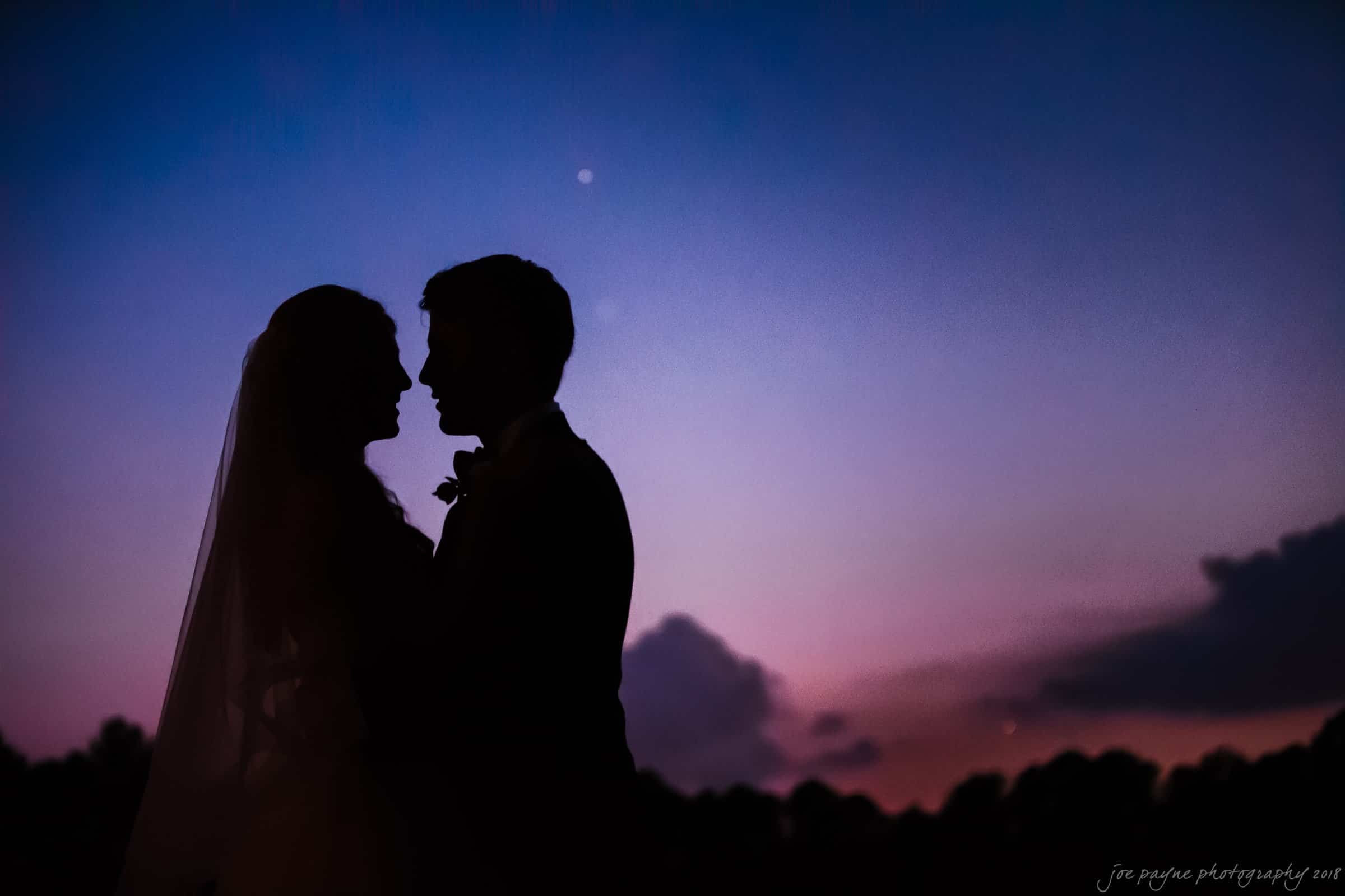 Silhouette of a bride and groom embracing against a vibrant sunset sky, with hues of pink, purple, and blue, and a single star visible.