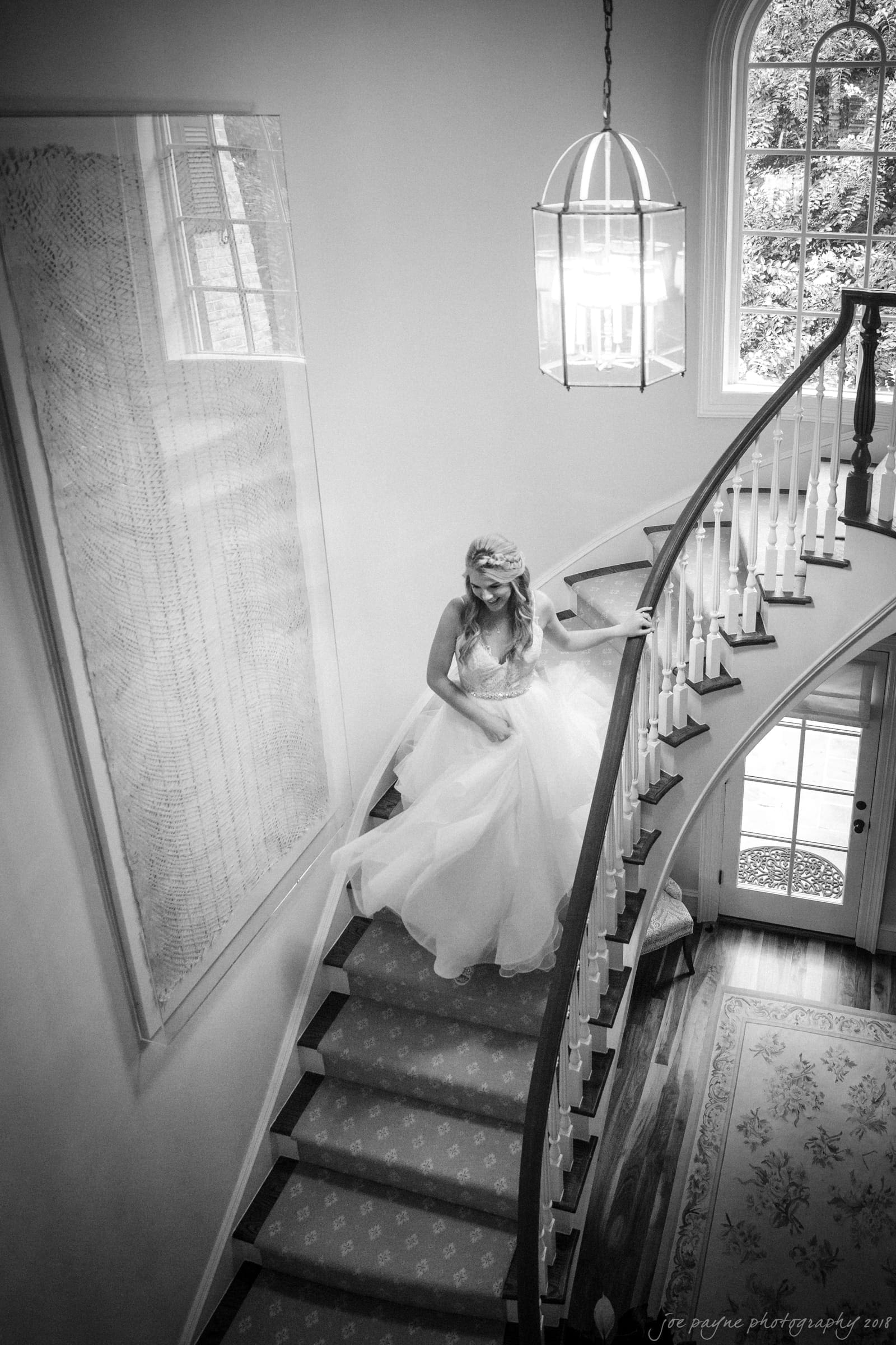 A bride in a flowing gown descends a curved staircase with a large window in the background. The scene is captured in black and white, emphasizing the elegant and serene atmosphere.