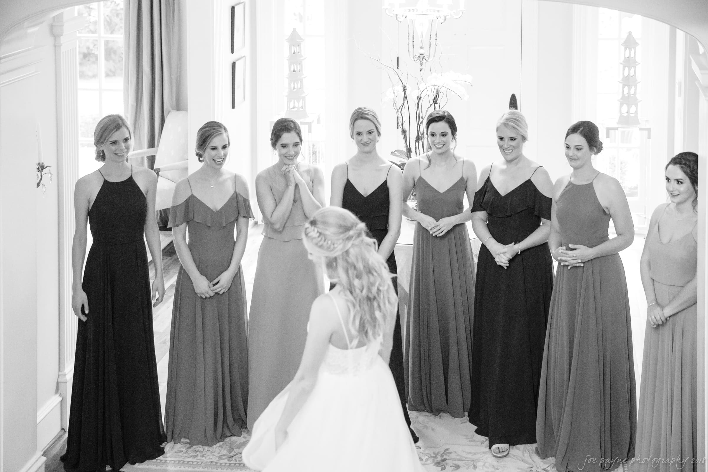 A bride in a white gown faces a group of bridesmaids wearing different styles of dark and light dresses. They are standing in a well-lit room with elegant decor and are smiling and looking at the bride.
