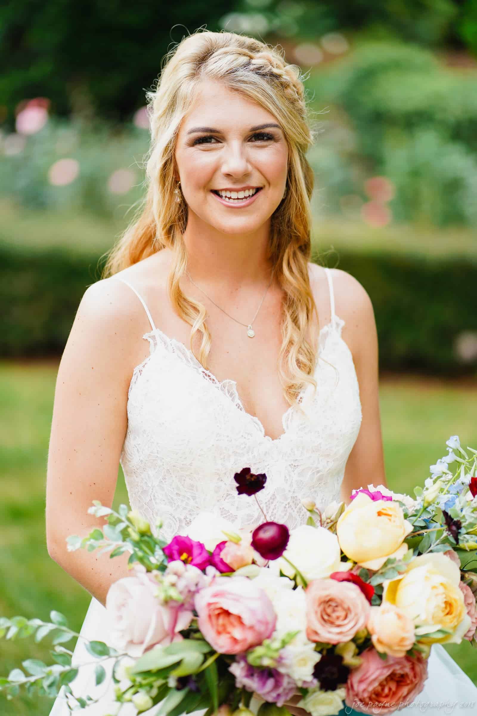A smiling bride in a white lace dress holds a colorful bouquet with roses and peonies in an outdoor garden setting. Her blonde hair is styled in loose waves.