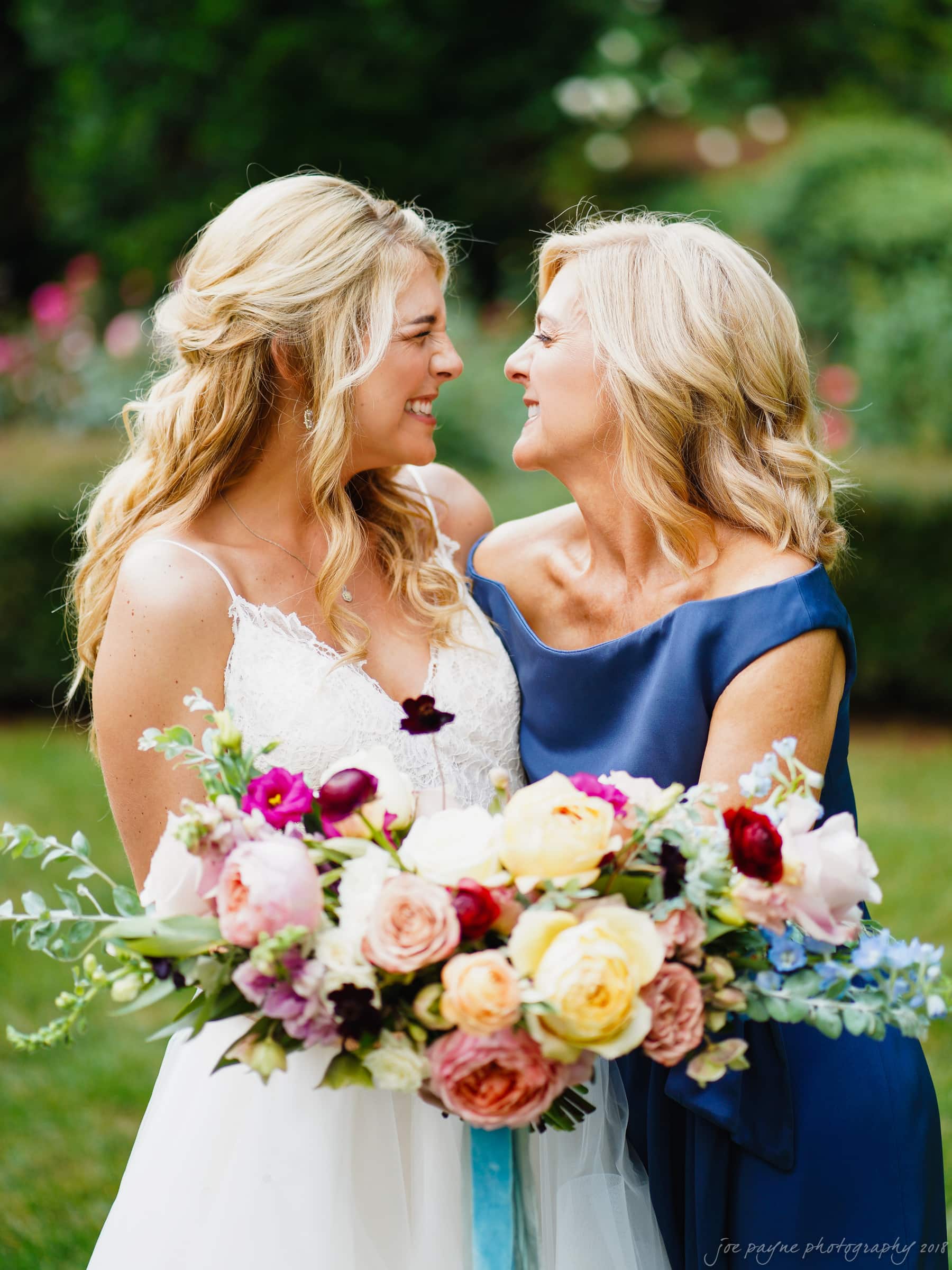 Two women smiling at each other in an outdoor setting. One is in a white wedding dress and the other in a blue dress. They are holding a large, colorful bouquet of flowers. Lush greenery is visible in the background.