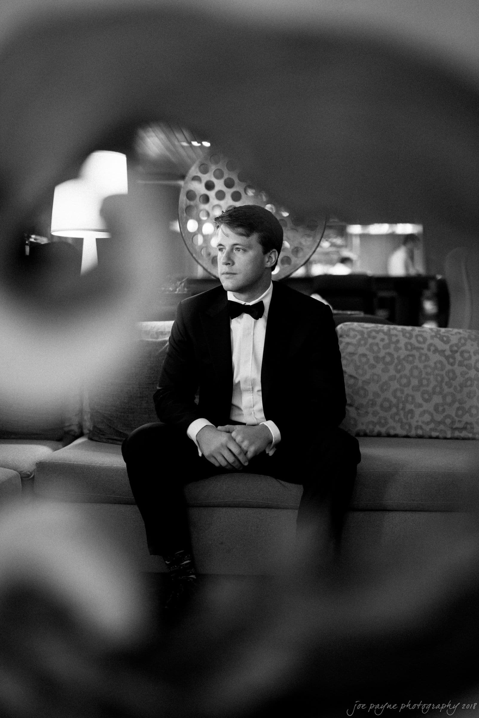 A man in a tuxedo sits thoughtfully on a couch, framed by a decorative element in the foreground. The black and white image conveys a classic and elegant atmosphere.