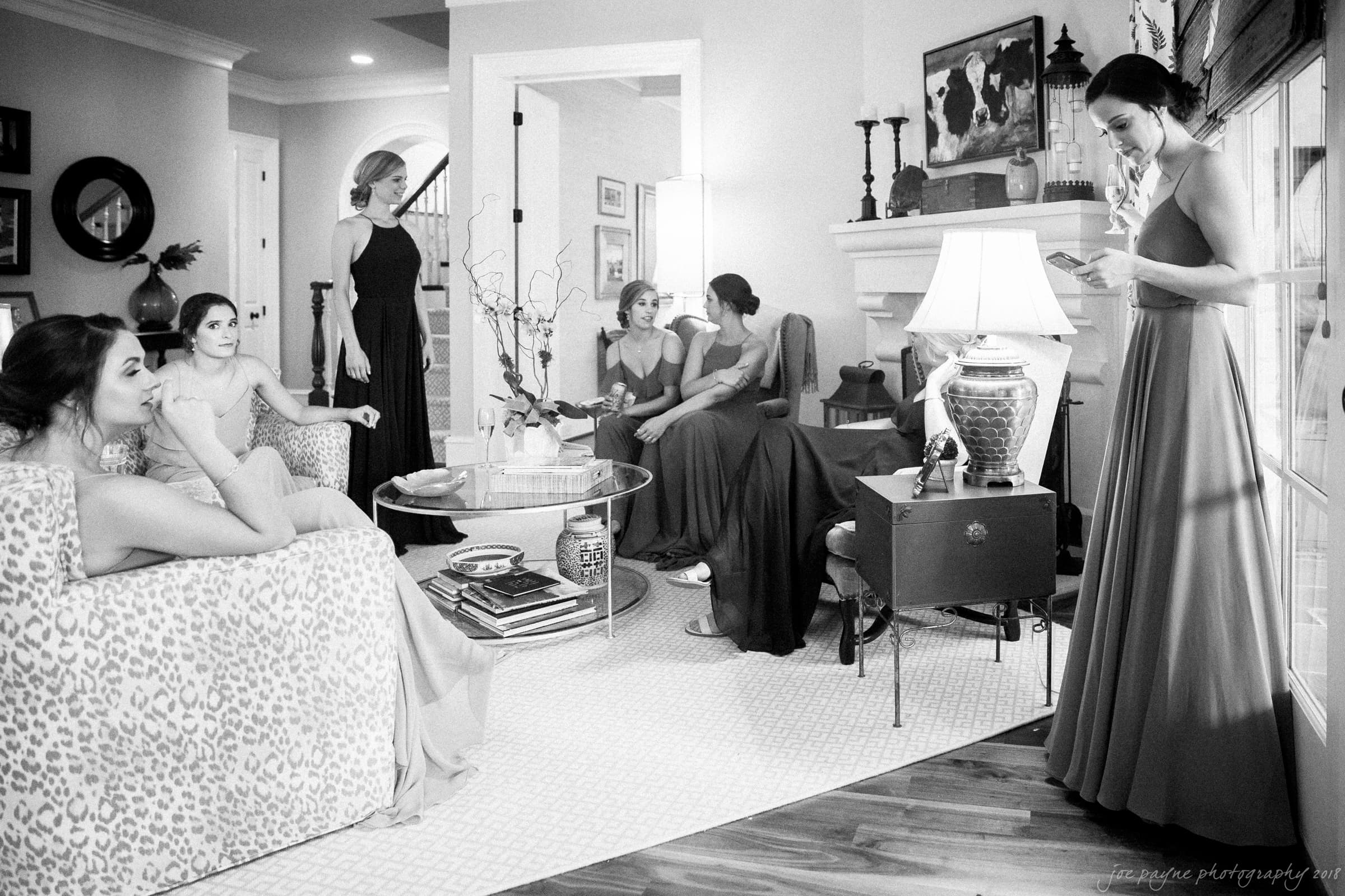 A black and white image of six women in elegant dresses gathered in a living room. They are engaged in different activities, such as reading and conversing. The room is stylishly furnished with a sofa, armchairs, and a lamp.
