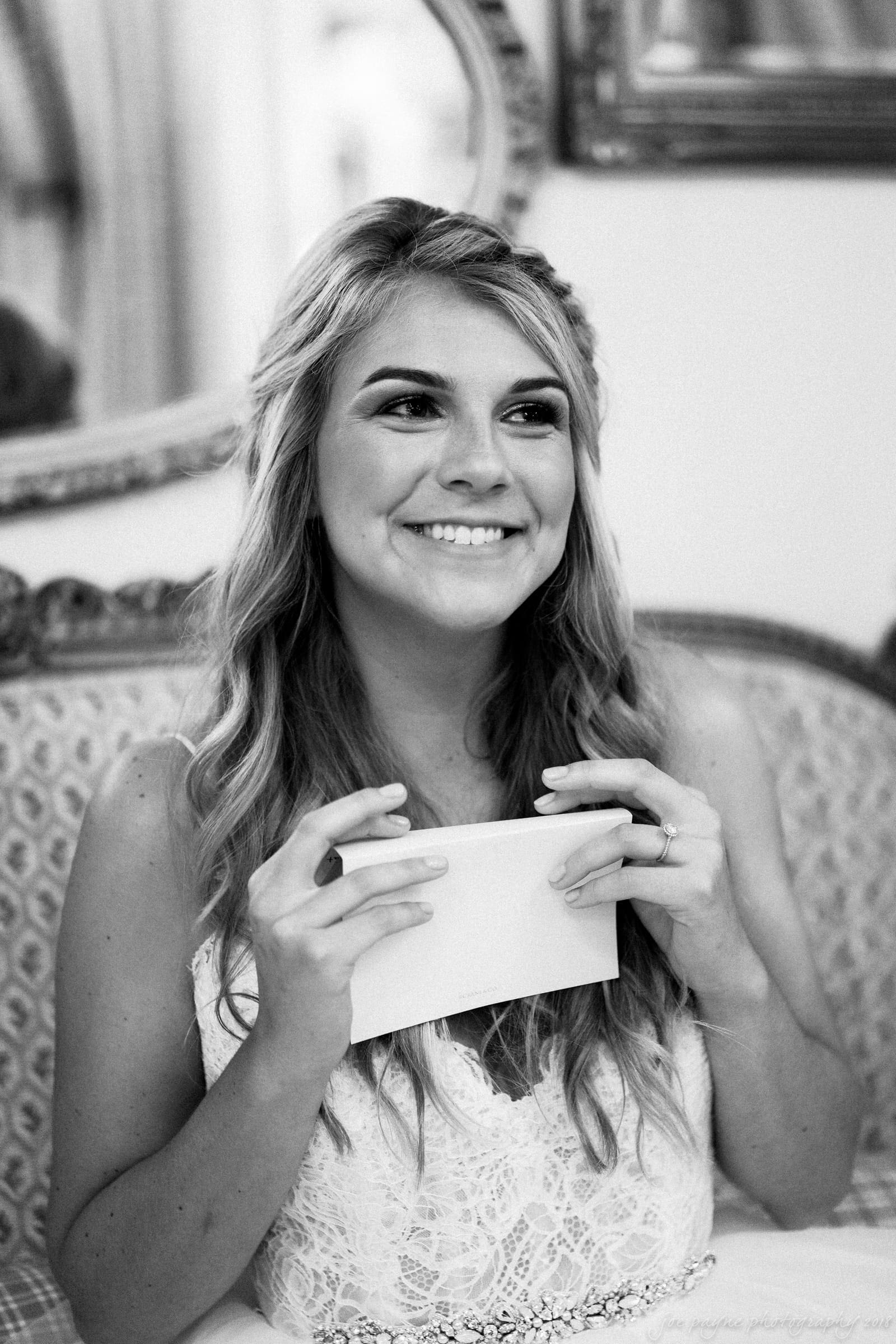 A woman with long hair sits on a patterned sofa, smiling while holding a piece of paper. She wears a white lace top, and the background features ornate mirrors. The setting appears to be indoors, possibly a living room. The image is in black and white.