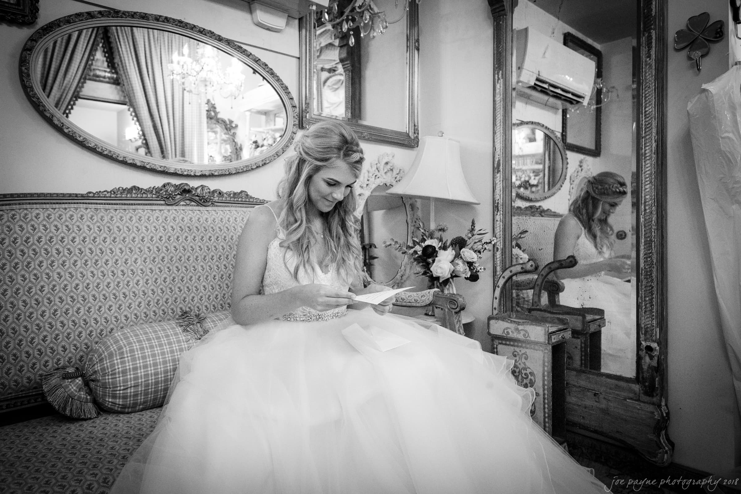 A bride in a white gown sits on a patterned sofa inside a vintage-style room. She is smiling while reading a letter. The room has ornate mirrors, a floral lamp, and curtains in the background.