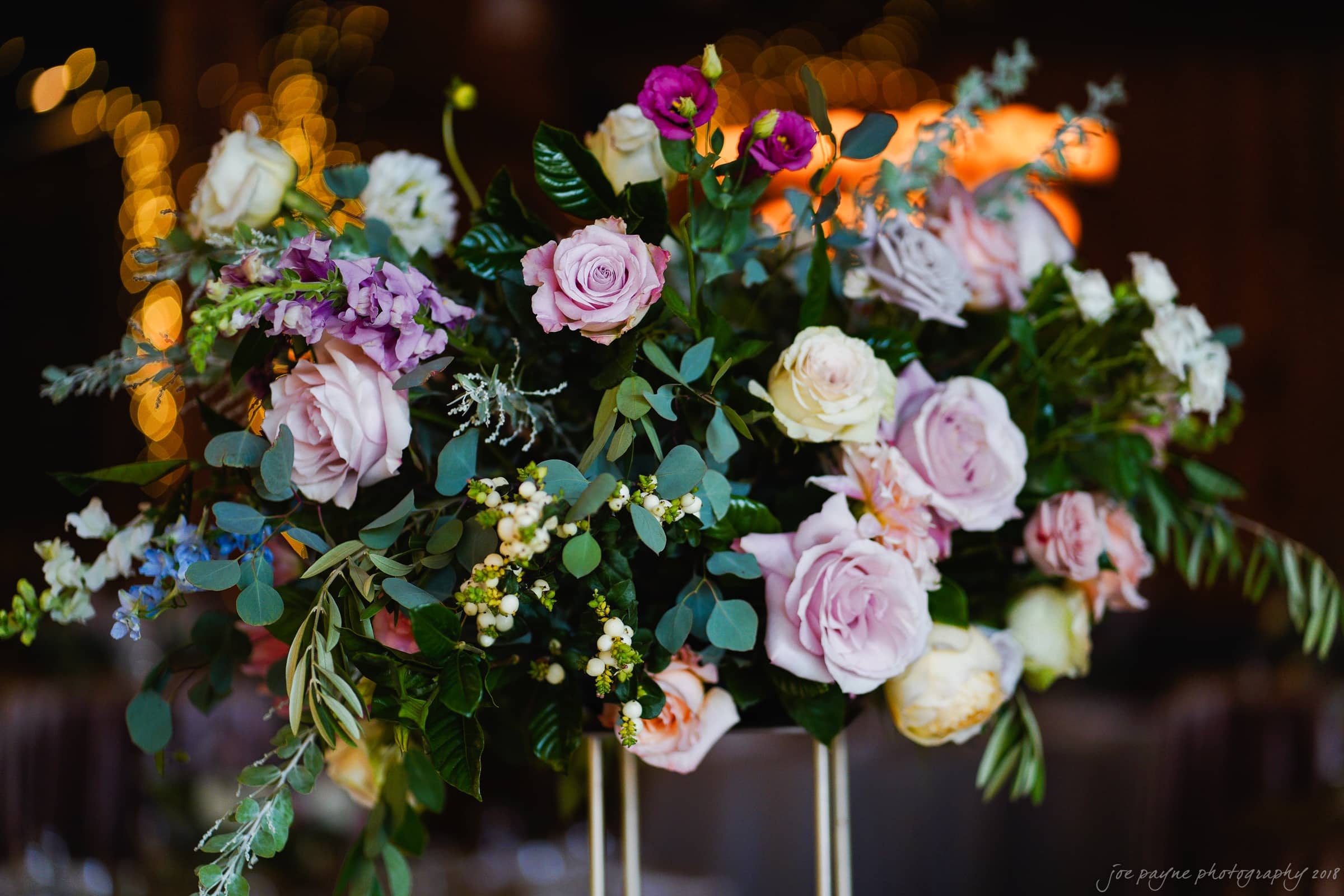A lush floral arrangement featuring pink and white roses, purple flowers, and various greenery, set against a softly lit background.